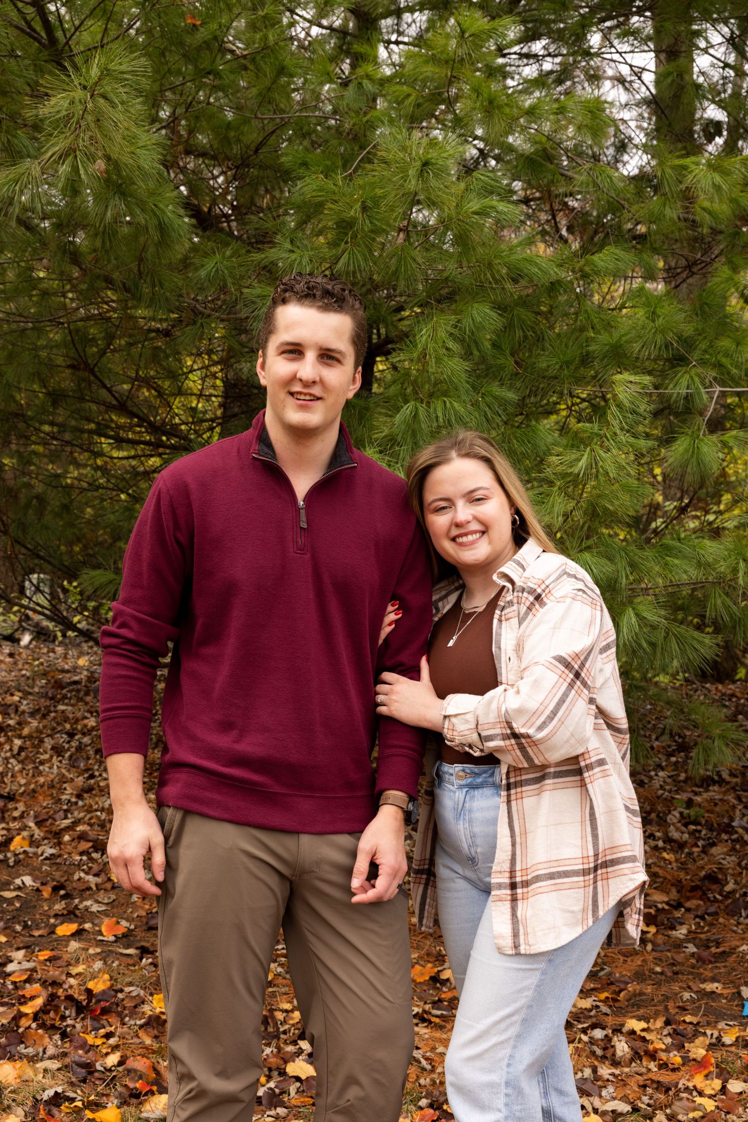 A couple poses playfully among autumn leaves and pine trees.