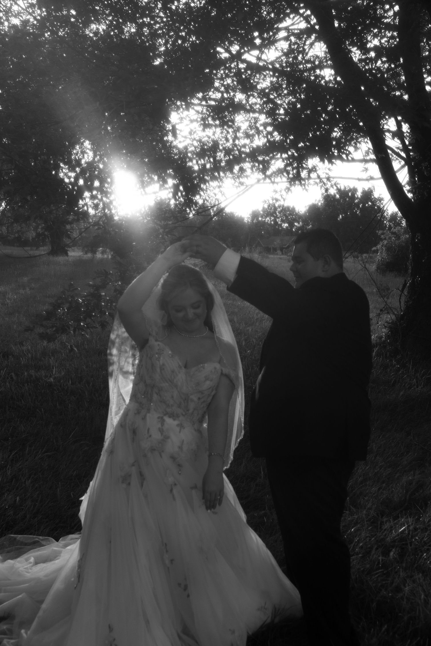 Black and white photo of a bride and groom dancing outdoors at sunset, with sun rays filtering through trees.