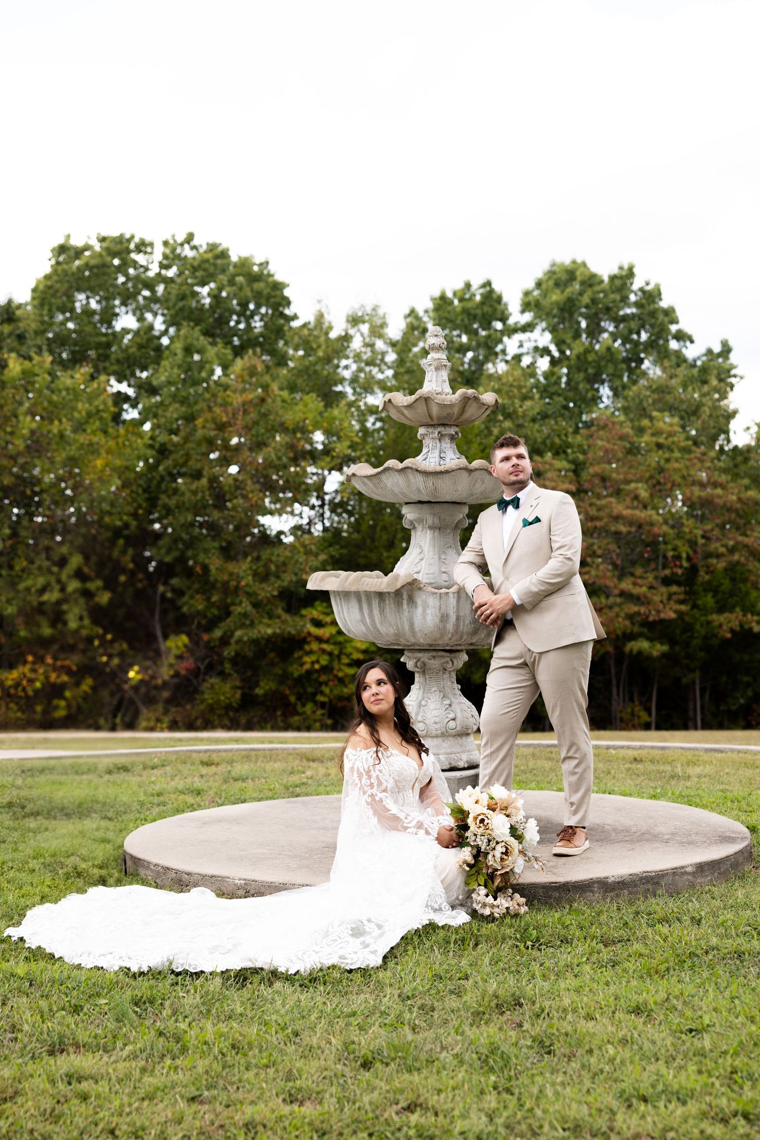 Wedding couple posing by a stone fountain in an elegant outdoor garden setting.