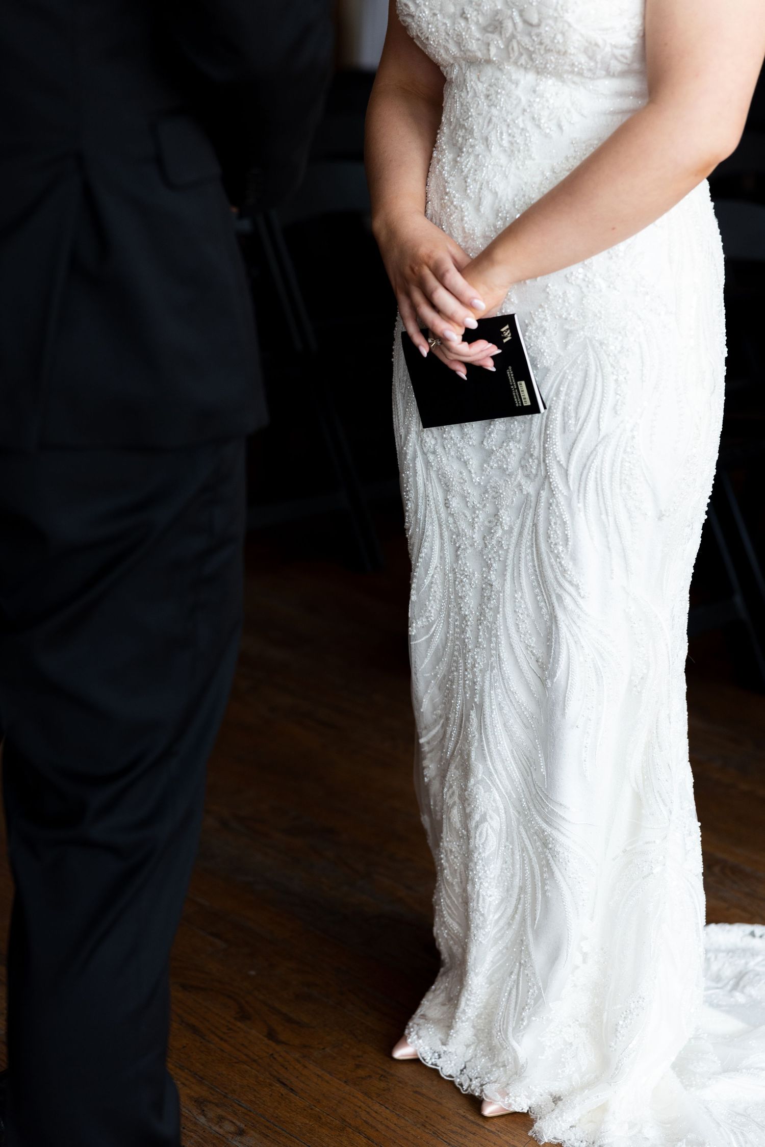 Bride in white lace wedding gown holding black clutch purse beside groom in black suit on wooden floor.