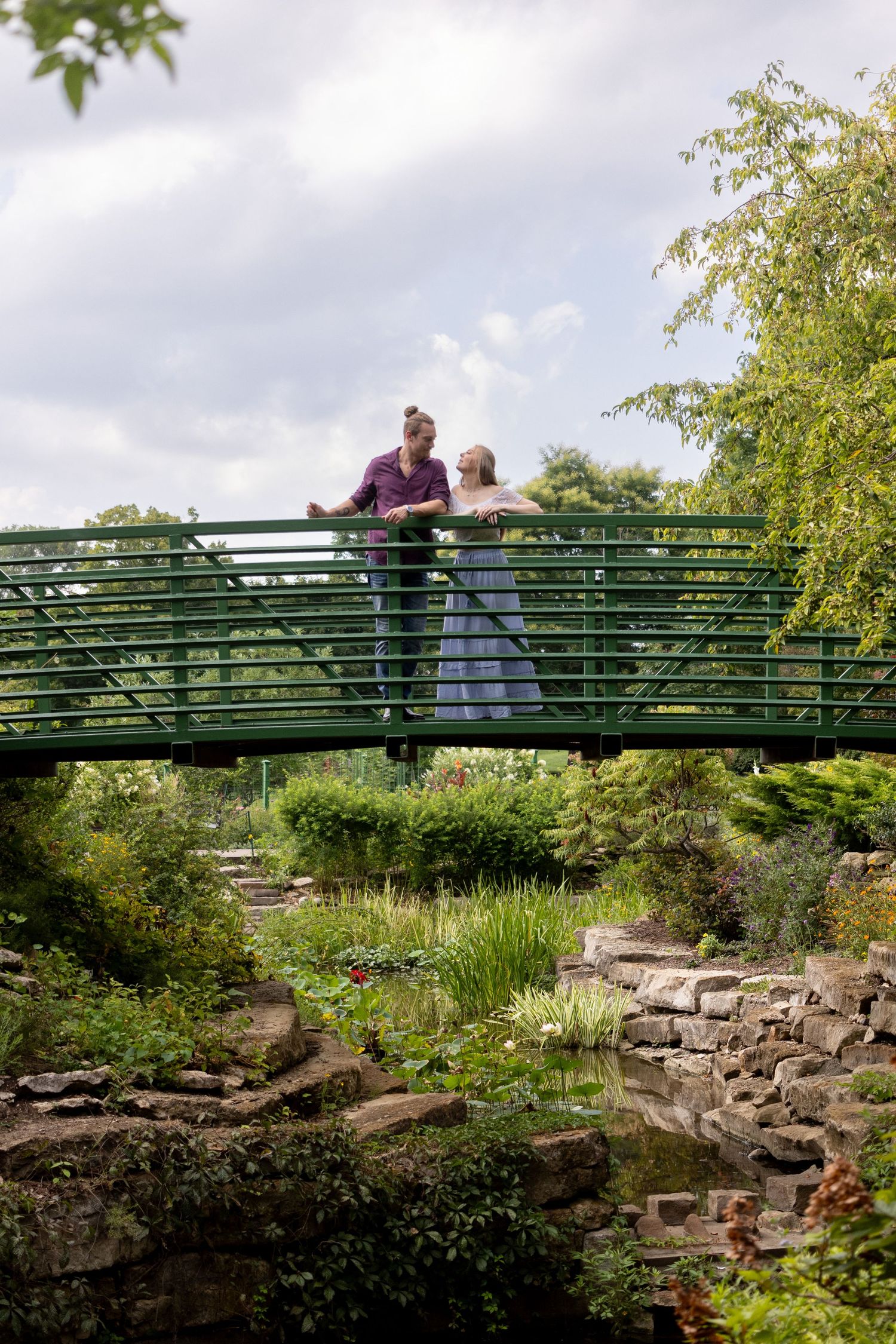 A couple stands on a green metal footbridge overlooking a garden with stone walls and lush vegetation.