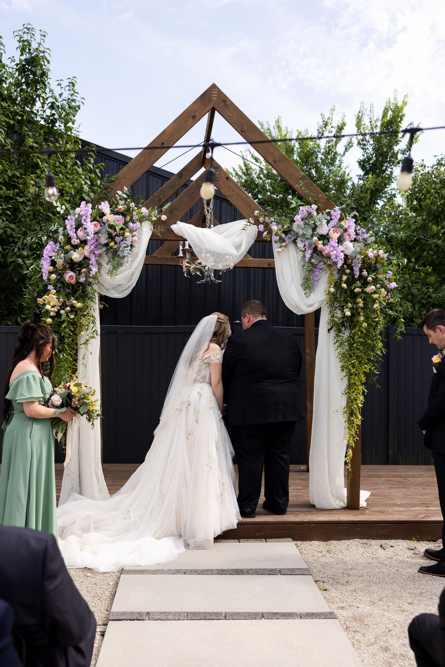 Bride and groom stand under a floral wooden arch with white draping during an outdoor wedding ceremony.