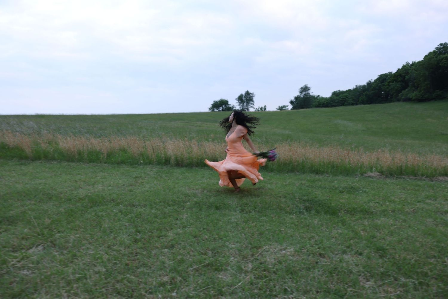 Woman in flowing orange dress spinning joyfully in a lush green field with trees in the background.