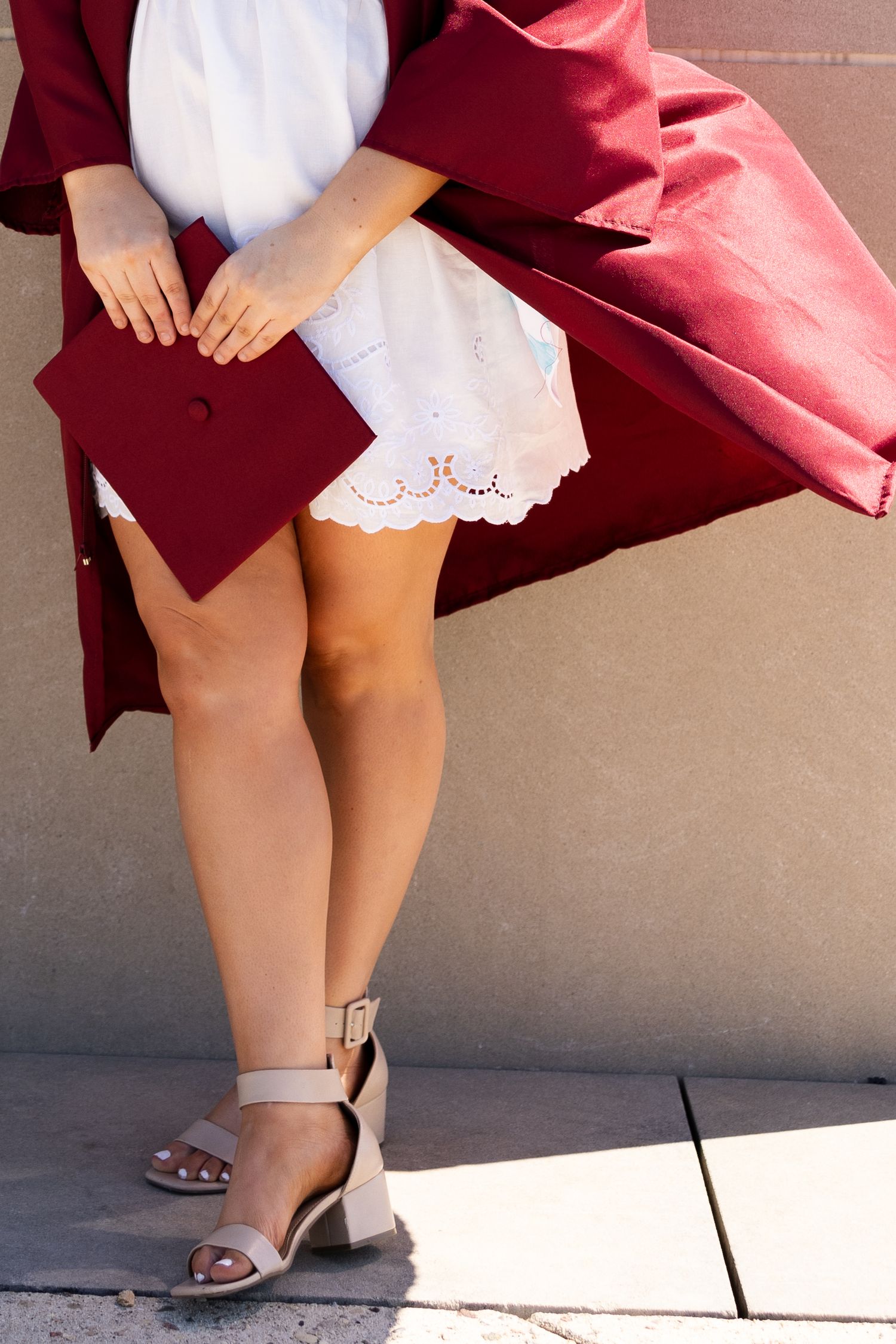 Graduate in red gown holding cap, wearing white dress and nude block heel sandals, standing outdoors.