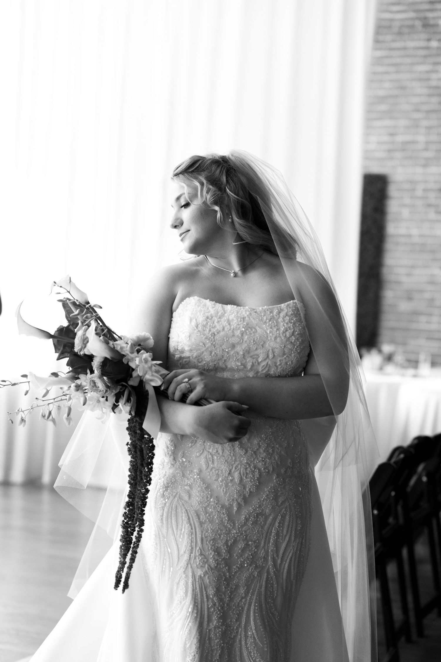 Black and white photo of a bride in lace gown and veil holding a floral bouquet, smiling softly indoors.