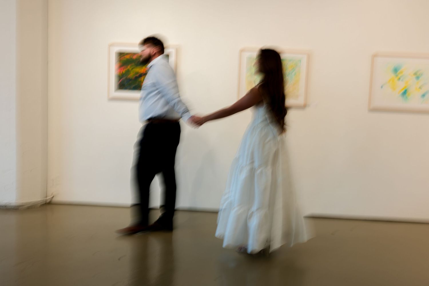 Motion blurred couple dancing in an art gallery with white walls and framed artwork.