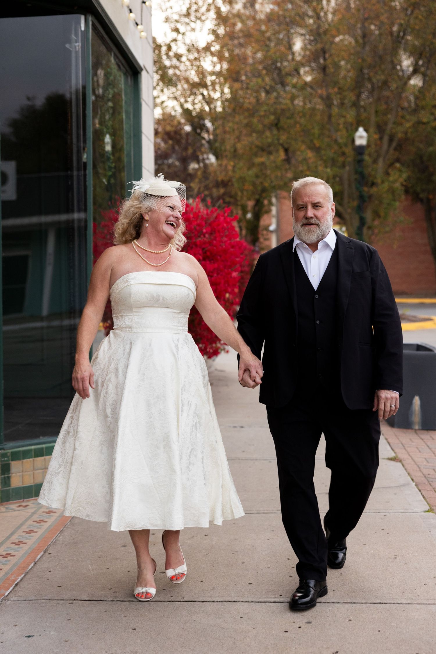 A couple walking together outdoors in formal wedding attire against a brick building backdrop.