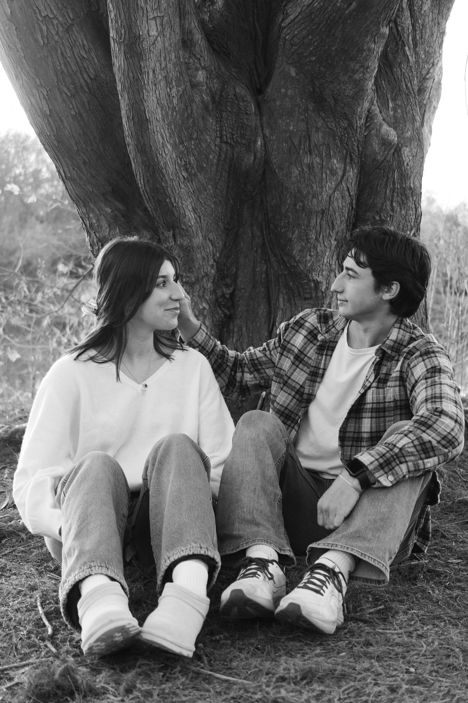 Couple sitting against large tree trunk sharing intimate moment in black and white photo.