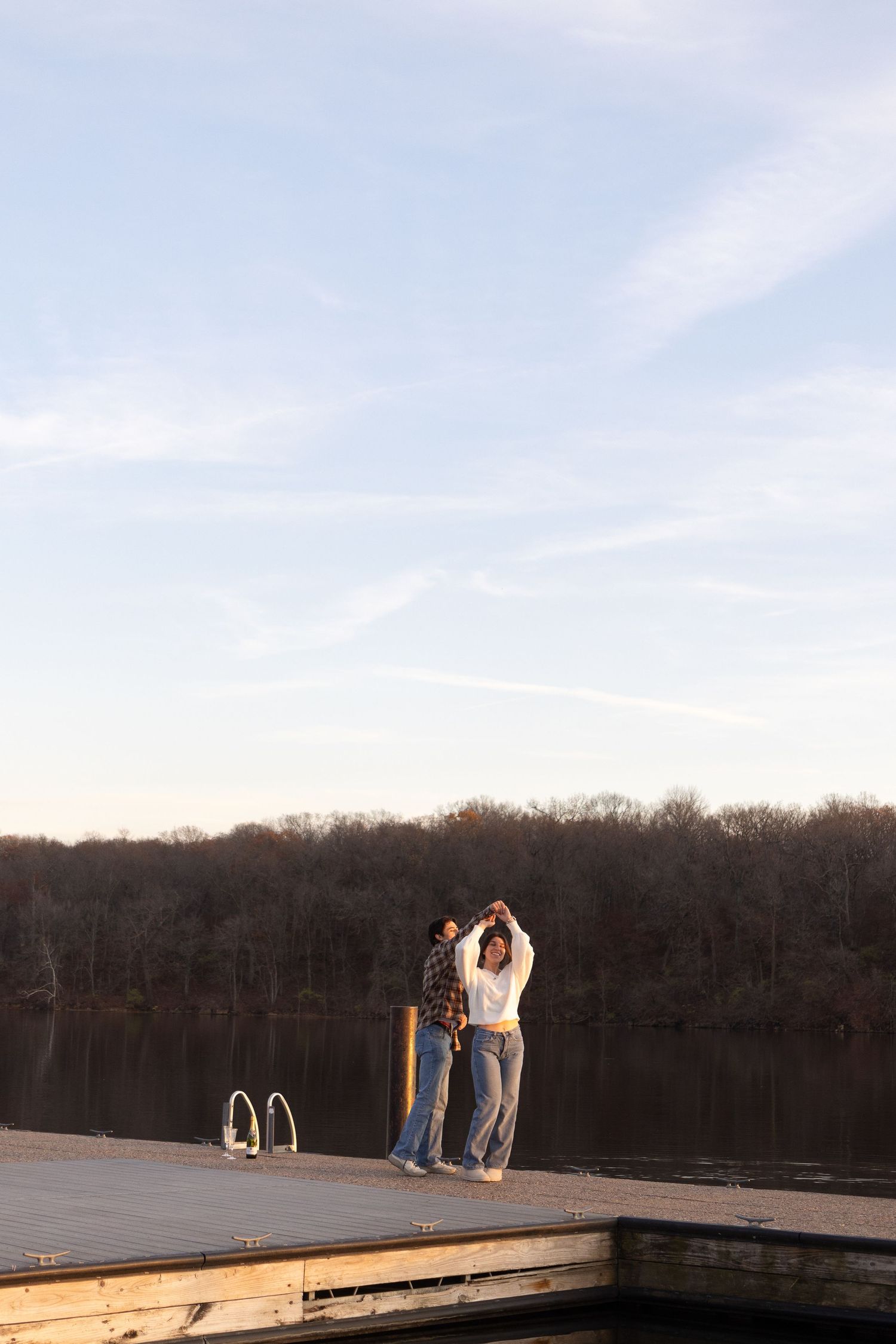 A couple standing on a wooden dock by a lake during sunset.