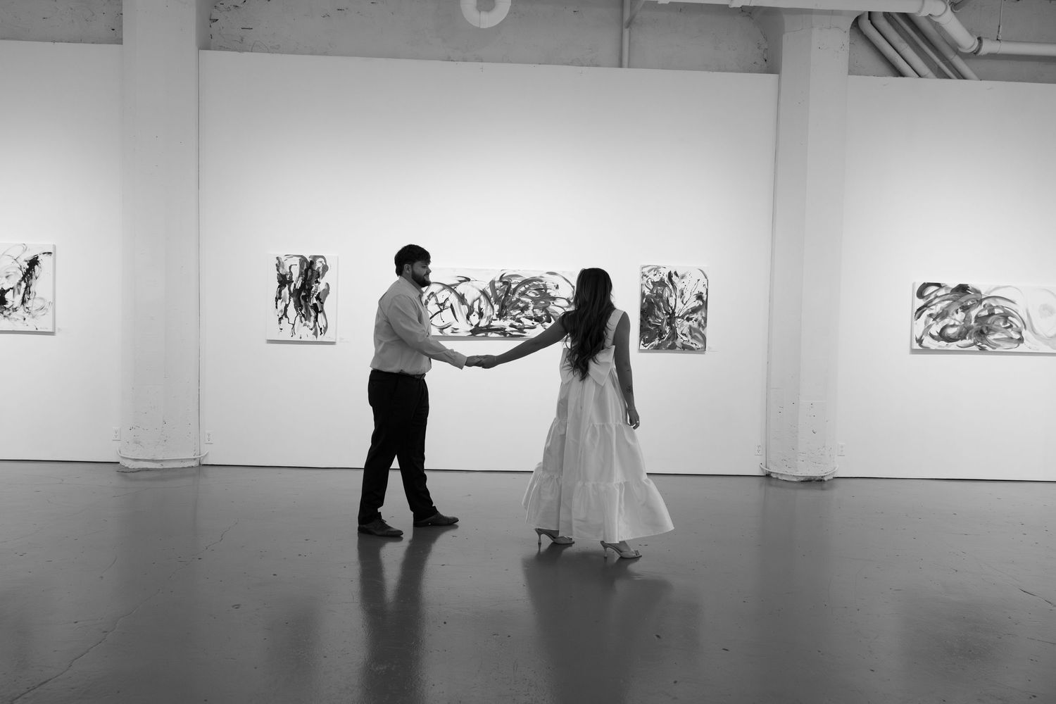 Black and white photo of a couple holding hands in an art gallery with expressive paintings on white walls.