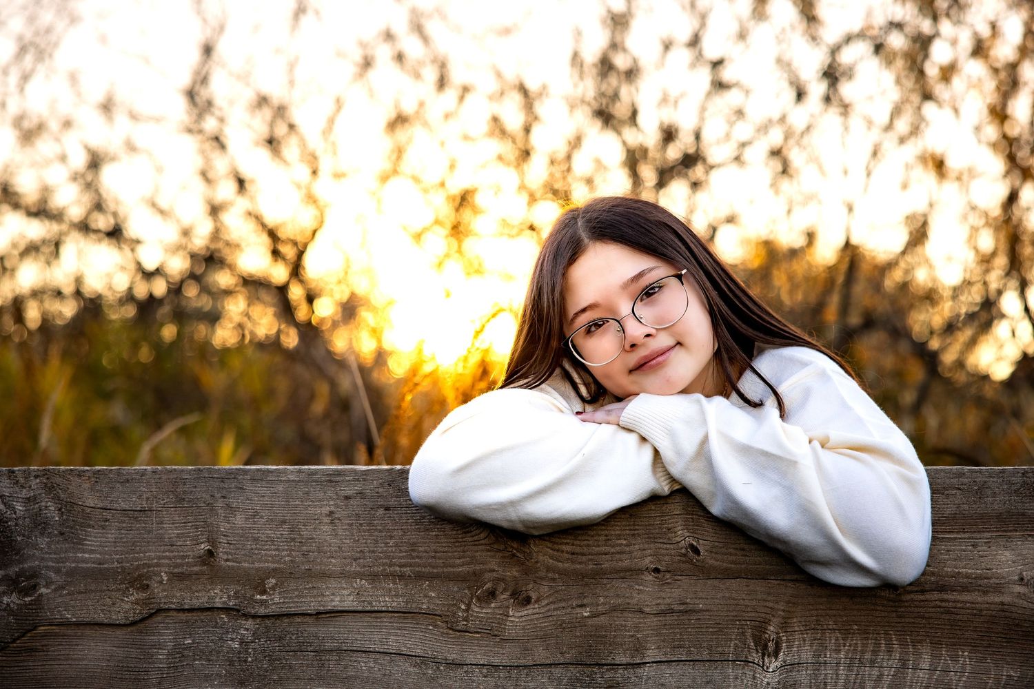 Person in a white sweater leans on wooden fence at sunset with golden sunlight filtering through trees in background.