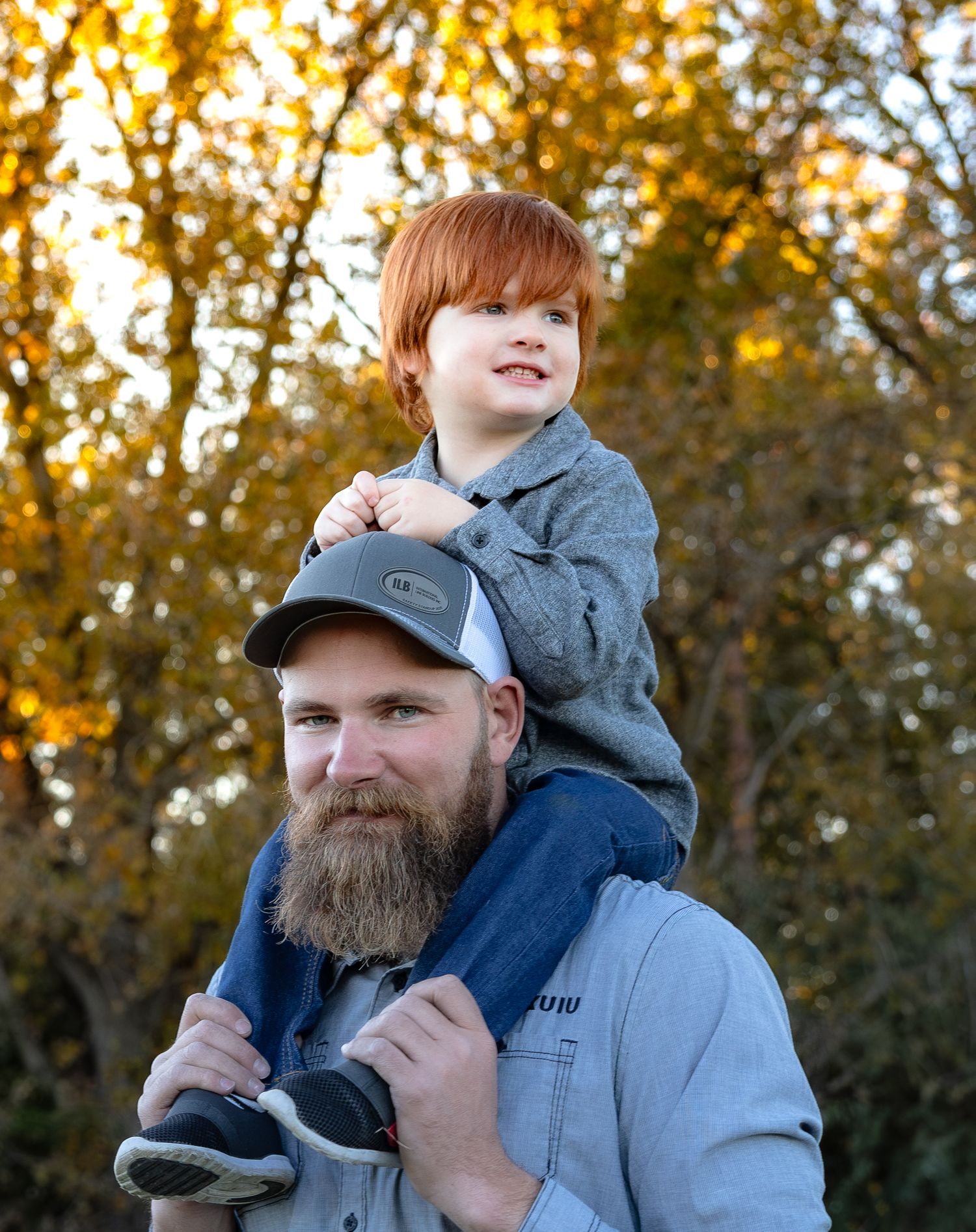 A person wearing a gray cap and blue jacket carries a child on their shoulders during a golden autumn afternoon.