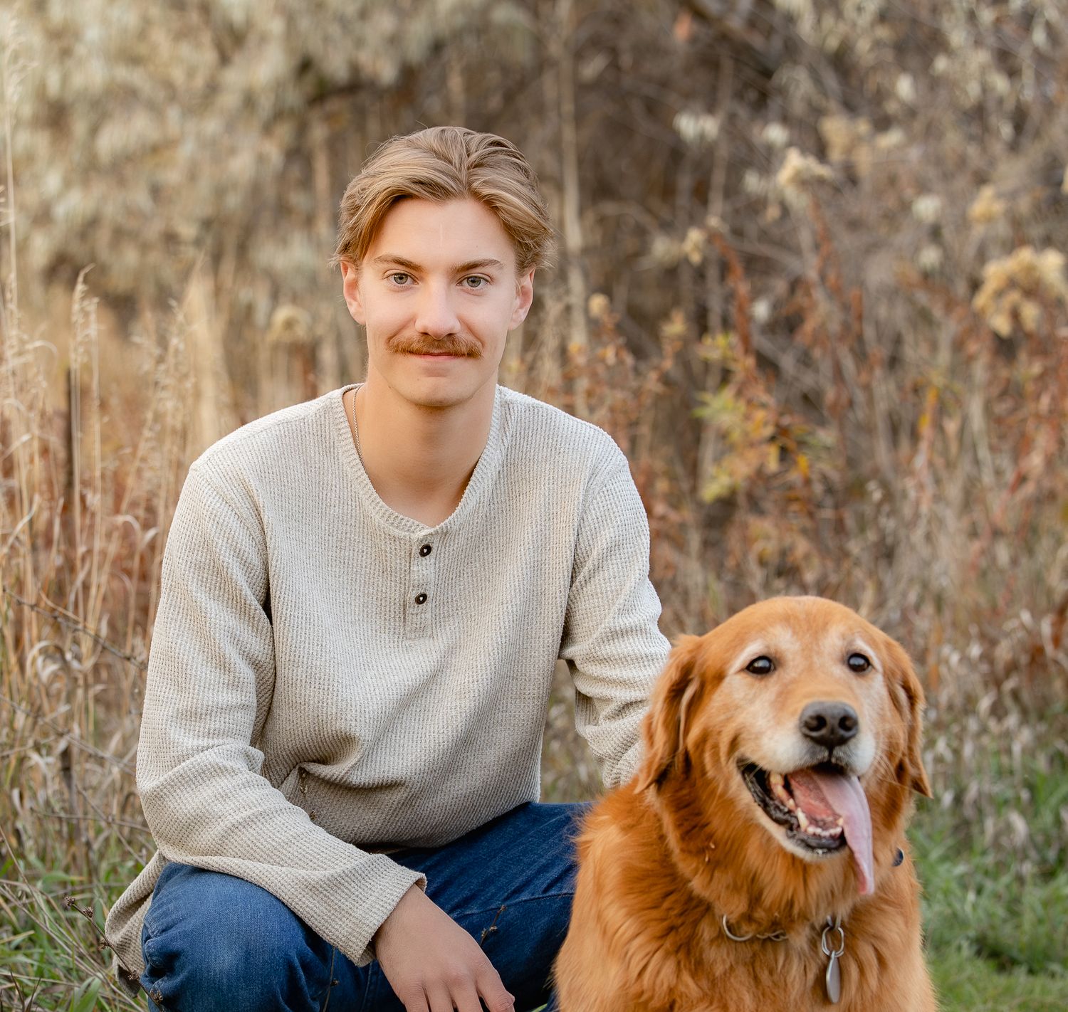 A golden retriever dog sits beside someone in a gray sweater during an autumn outdoor photoshoot.
