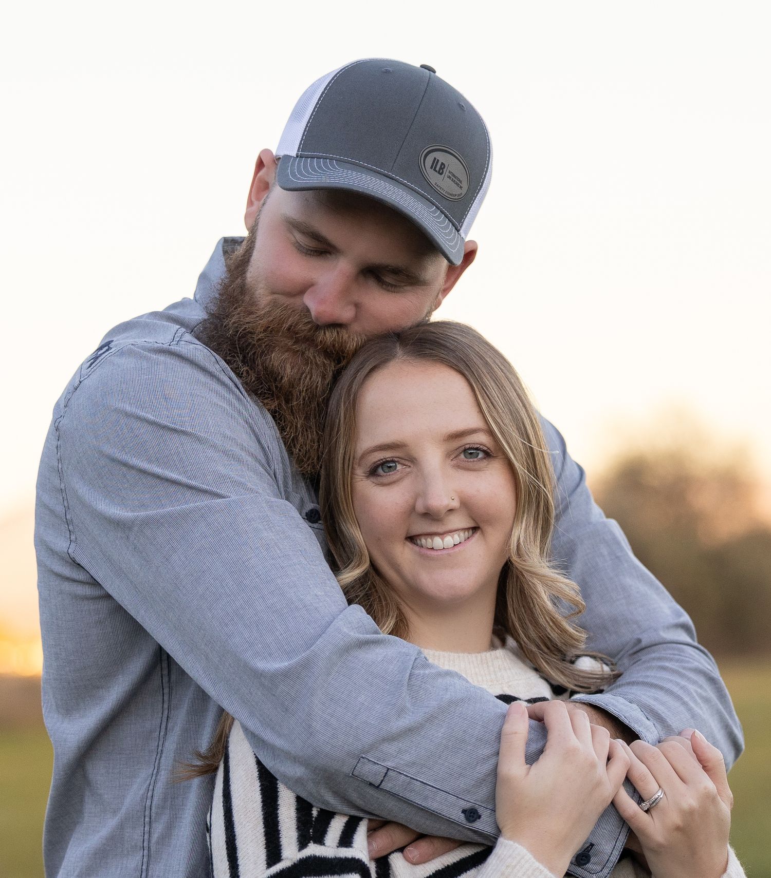 A couple embraces in a sweet moment during golden hour, with the person in a gray hat and sweater hugging from behind.
