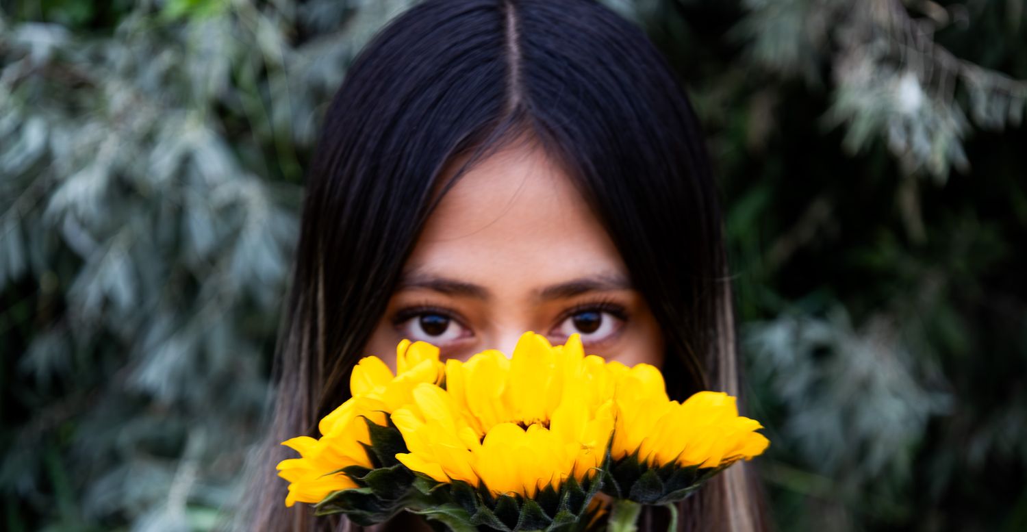 A woman peeks over bright yellow sunflowers in a creative outdoor portrait composition.