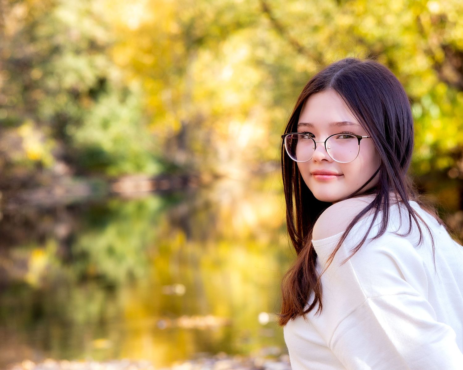 A person in a white shirt and glasses poses outdoors with autumn foliage and reflective water in the background.