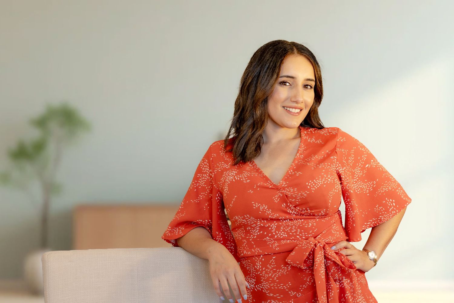 A person wearing a coral lace dress sits casually on a white couch in a bright, airy room.
