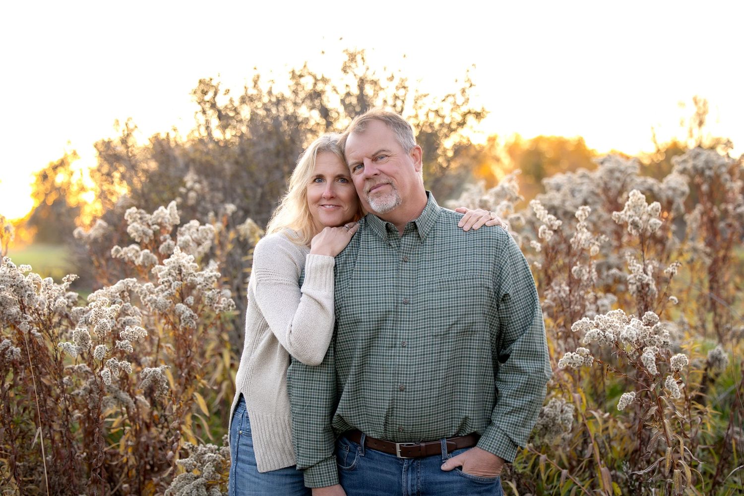 Couple embraces while standing in a sunlit field of tall grass during golden hour.