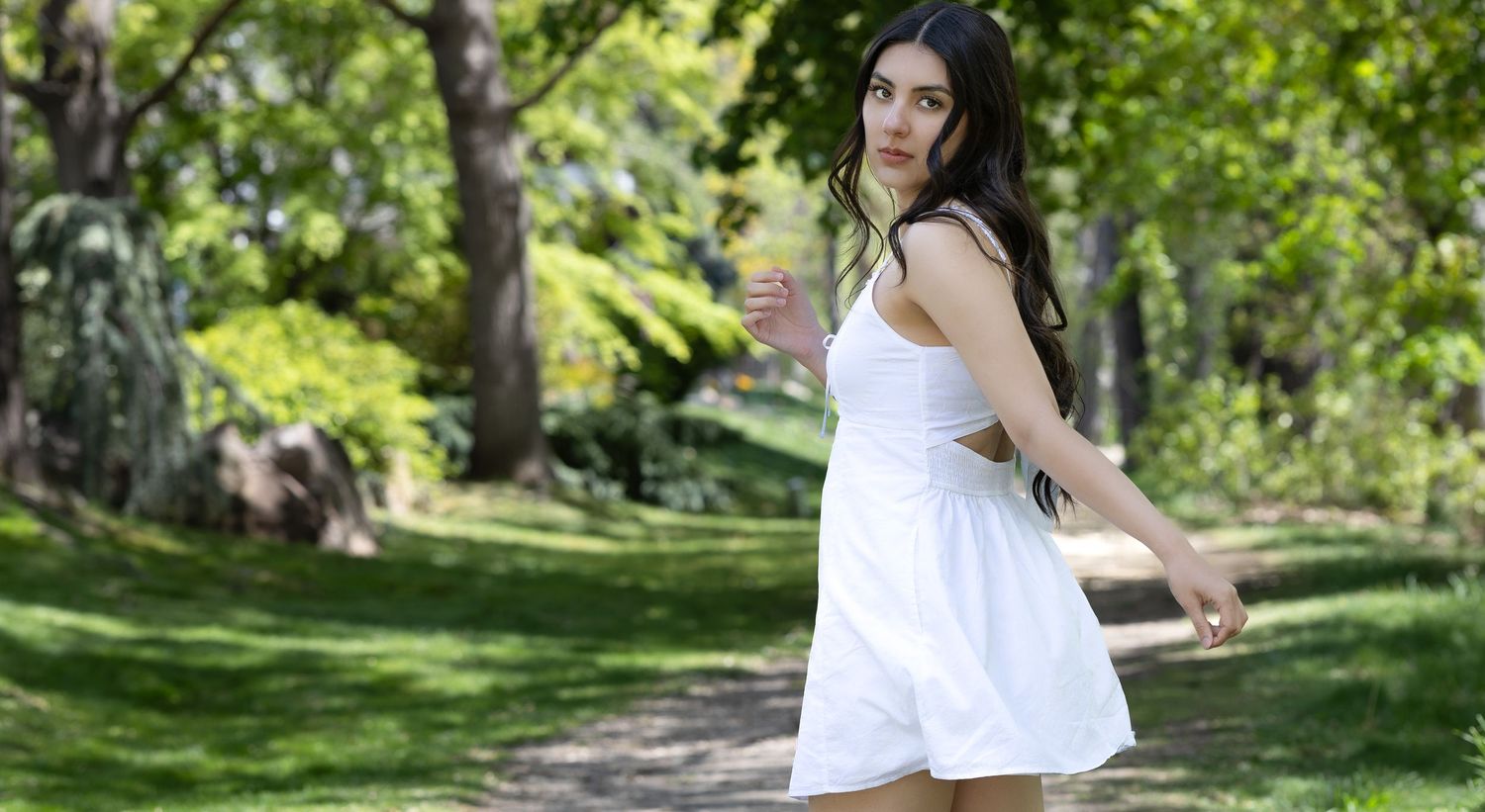Young woman in white sundress posing outdoors in lush green park on sunny day.