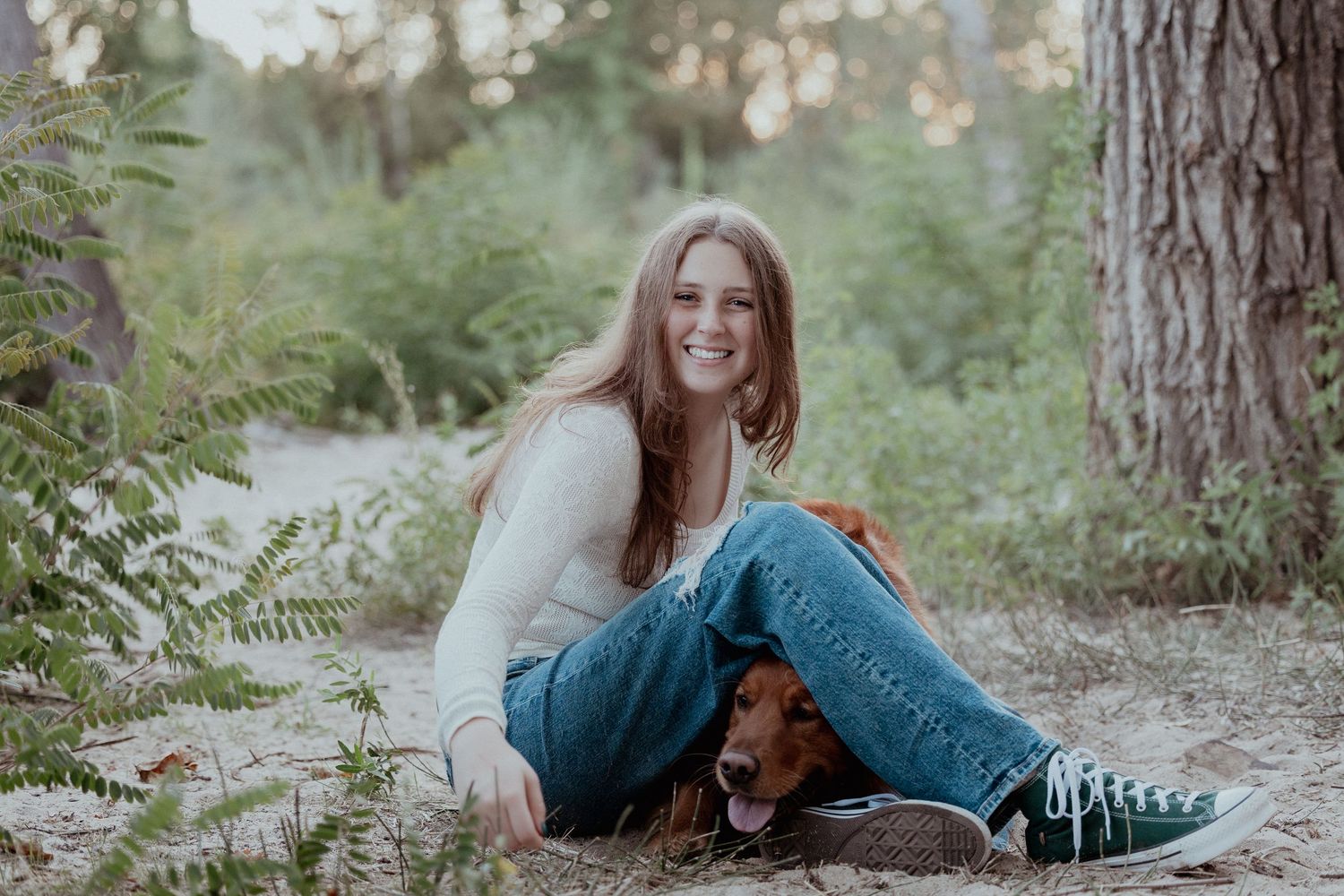 A person in jeans and teal sweater sits with a brown dachshund dog on a forest path surrounded by greenery.