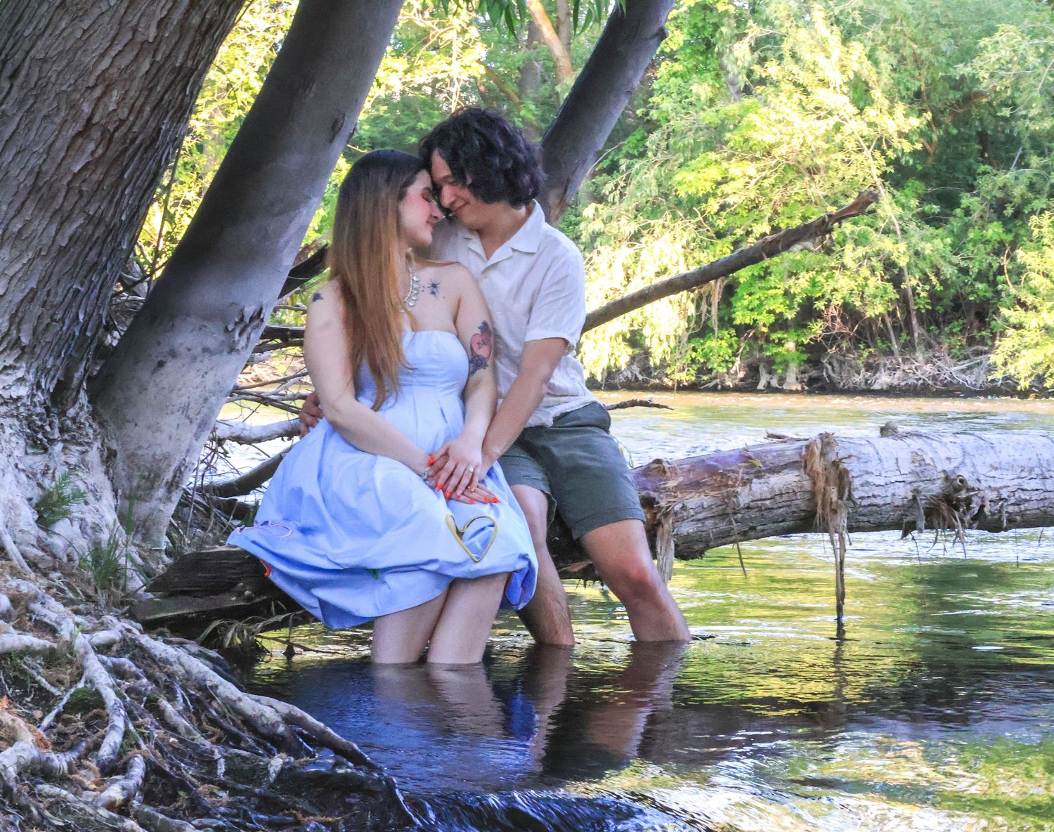 A couple in light summer attire sits together on a tree branch overhanging a peaceful river on a sunny afternoon.