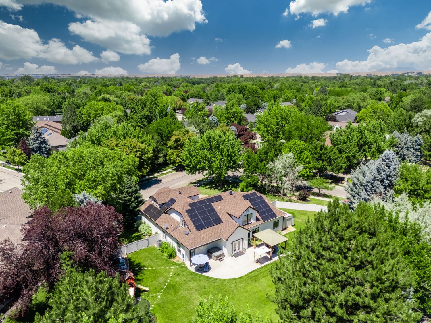 Aerial view of a suburban home with solar panels surrounded by lush green trees under a partly cloudy sky.