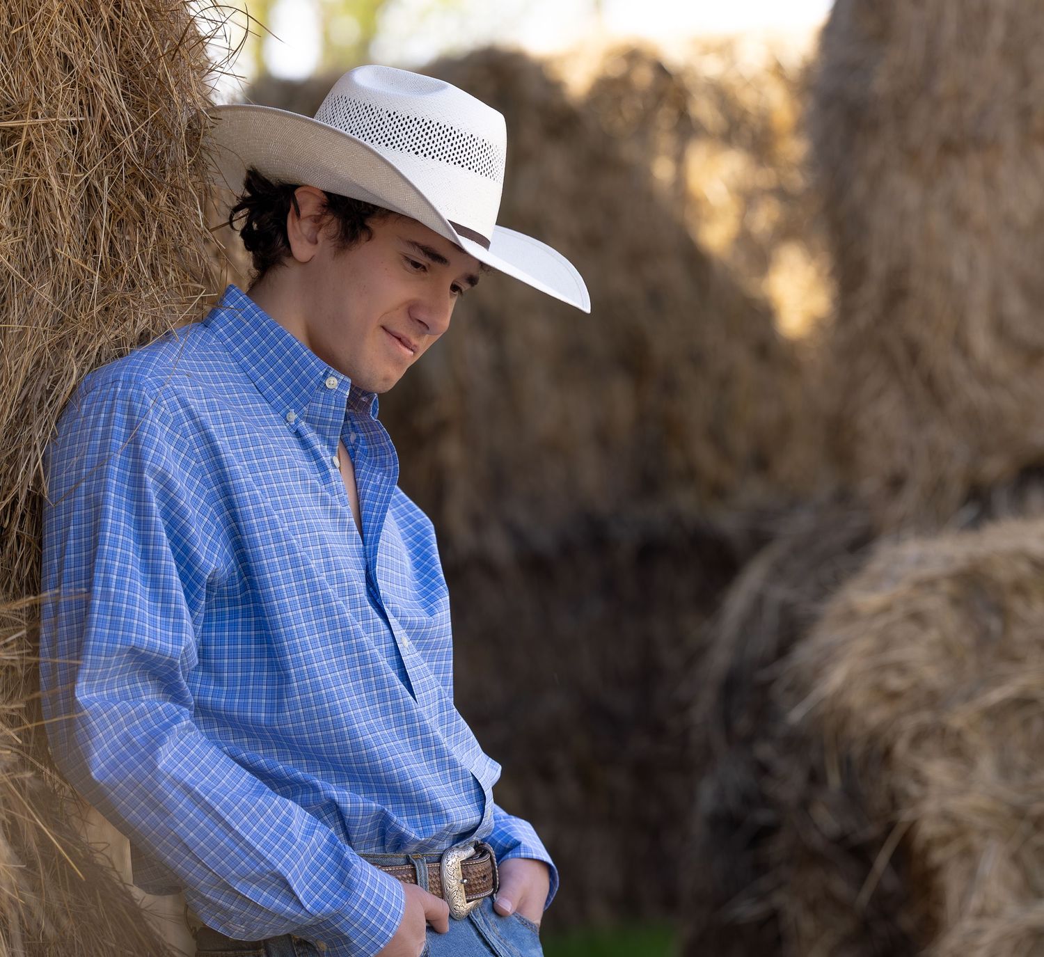 Young cowboy in white hat and blue plaid shirt leaning against hay bales in a rustic outdoor setting.