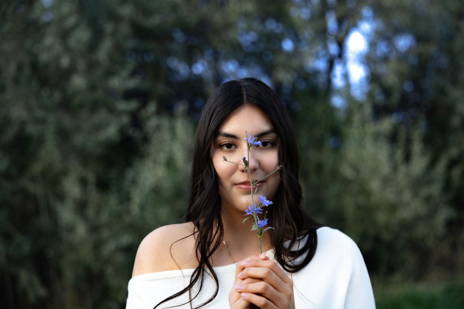 A person in a white off-shoulder top holds a small purple wildflower while standing in a lush outdoor setting.