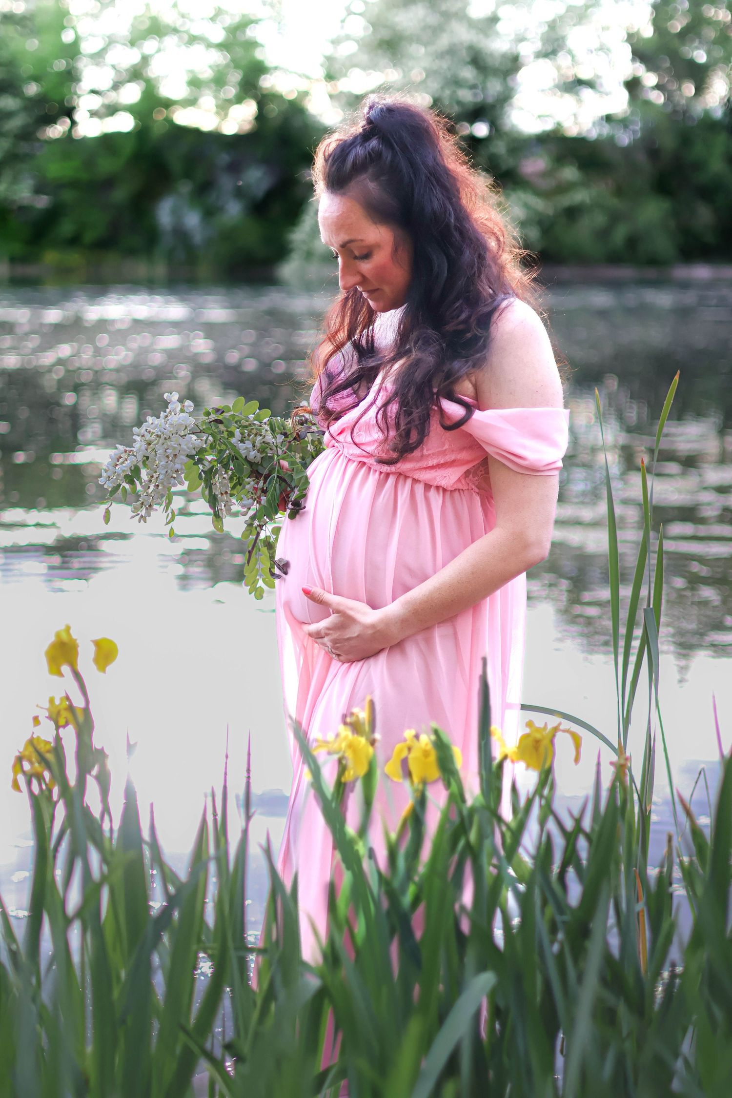 A pregnant woman in a pink dress stands among yellow iris flowers by a pond.