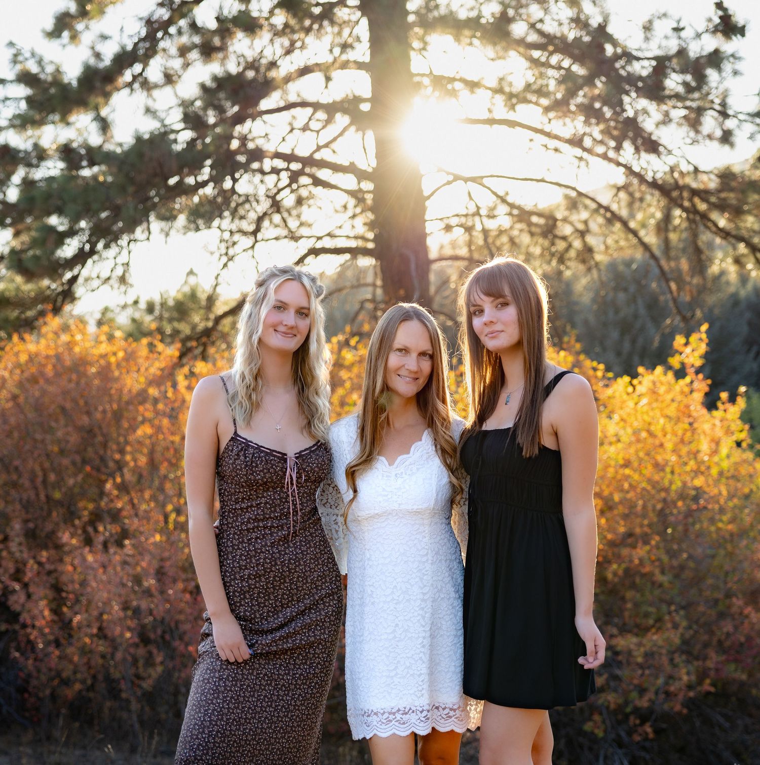 Three friends in casual outfits pose together at sunset with golden sunlight filtering through trees in the background.