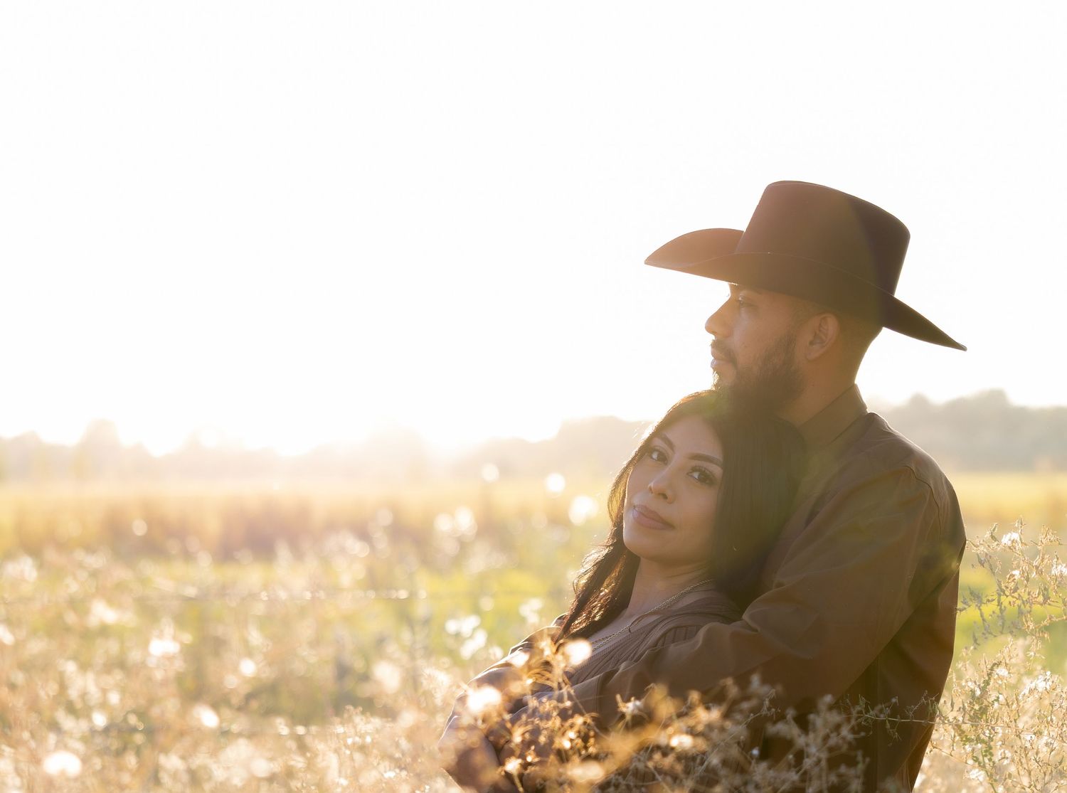 Romantic couple embraces in a sunlit meadow at golden hour with person wearing cowboy hat.