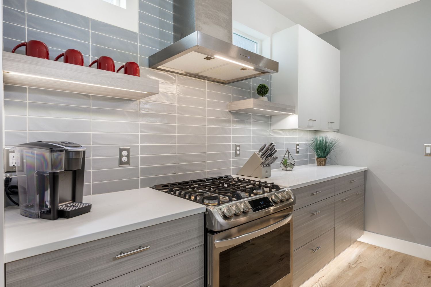 Modern kitchen with stainless steel gas range, gray tile backsplash, white countertops, open shelving, and light wood floors.