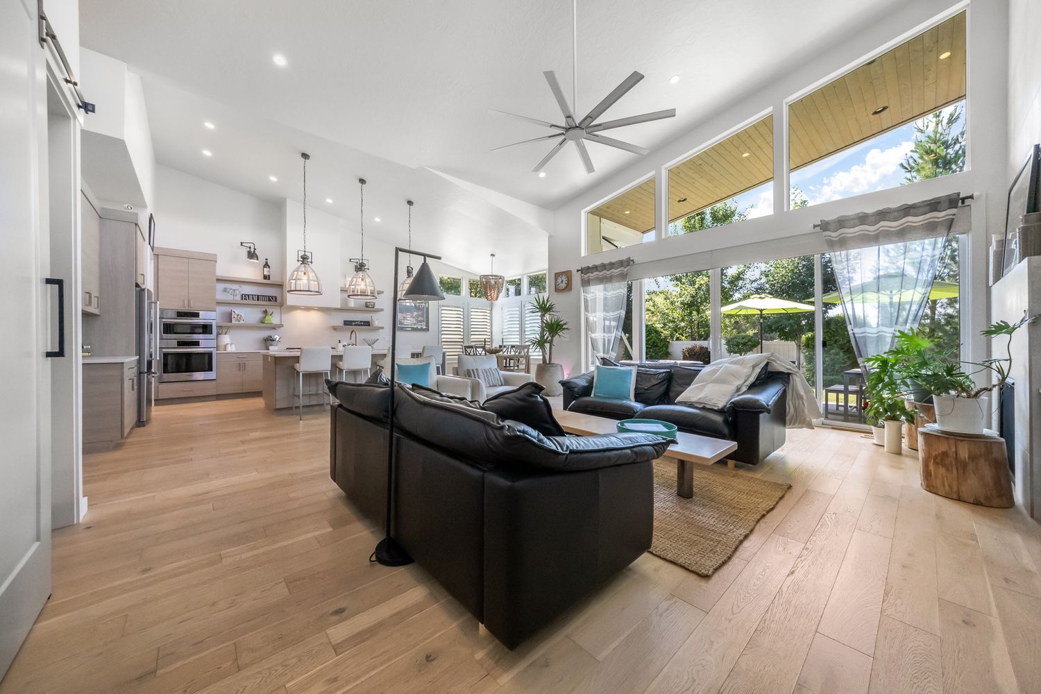 Modern open concept living room with hardwood floors, black leather sofa, and large windows overlooking a tropical garden.
