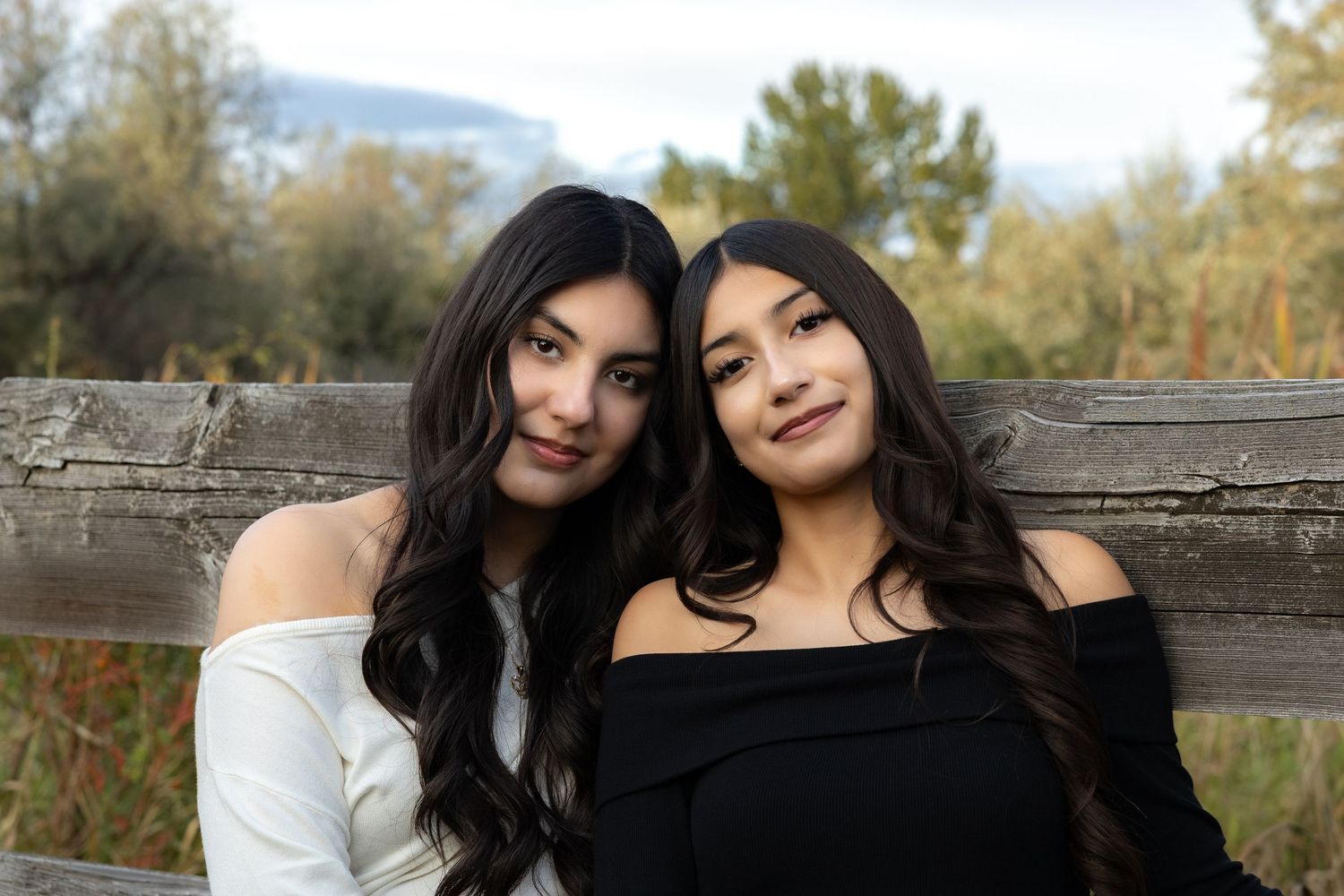 Two people sit close together on a wooden bench outdoors surrounded by autumn foliage and natural light.