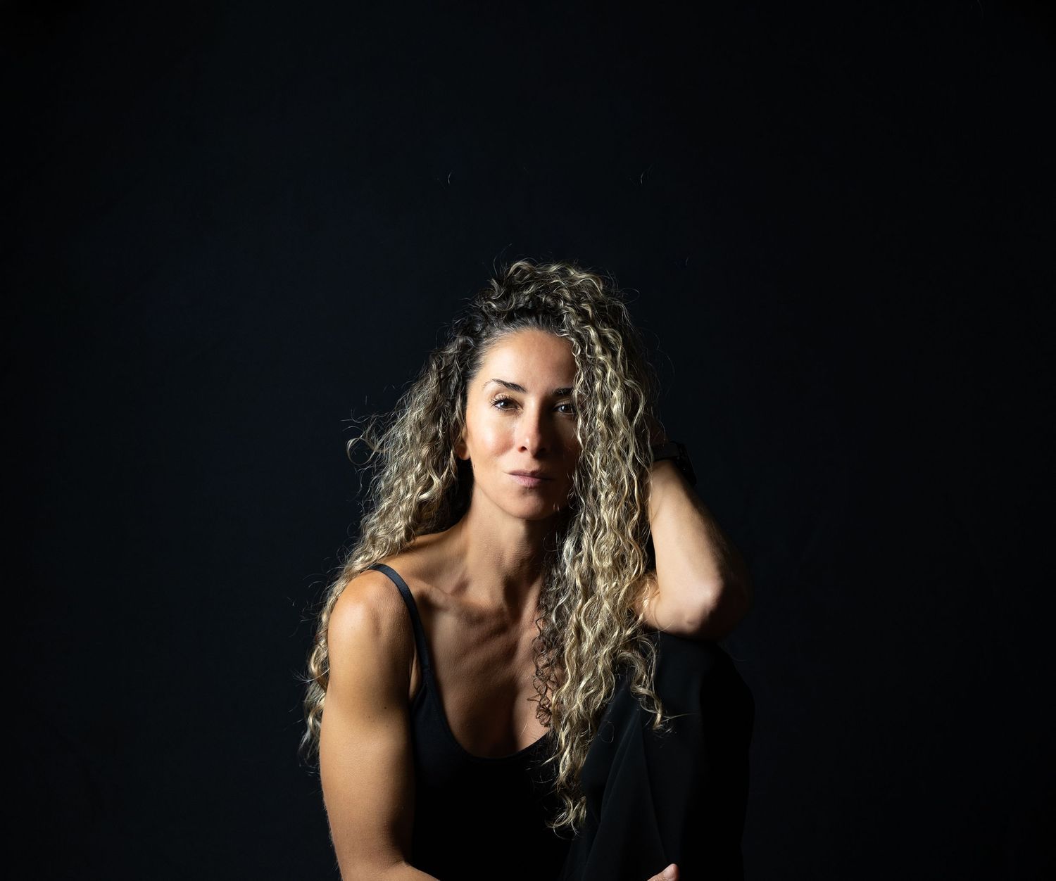 Athletic woman with long curly blonde hair poses against dark background in workout attire.