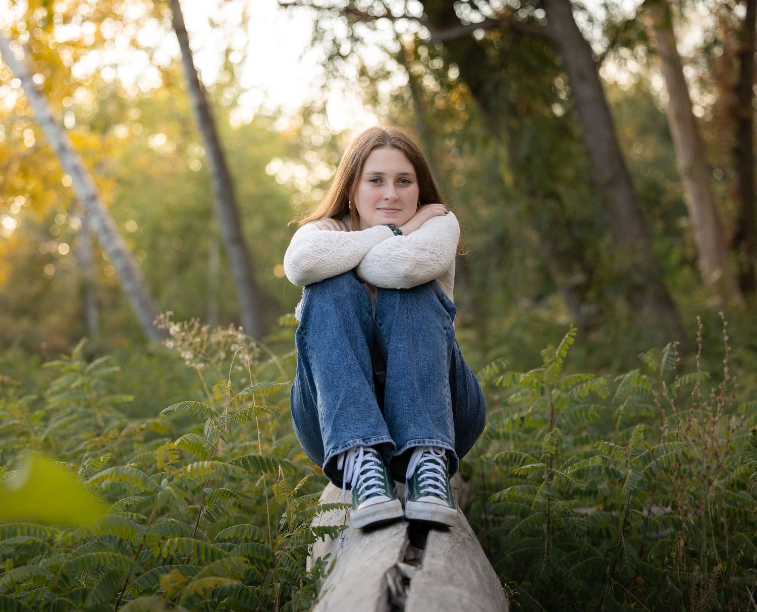 A person in a white dress and sneakers sits on a fallen tree trunk in a sunlit forest setting.