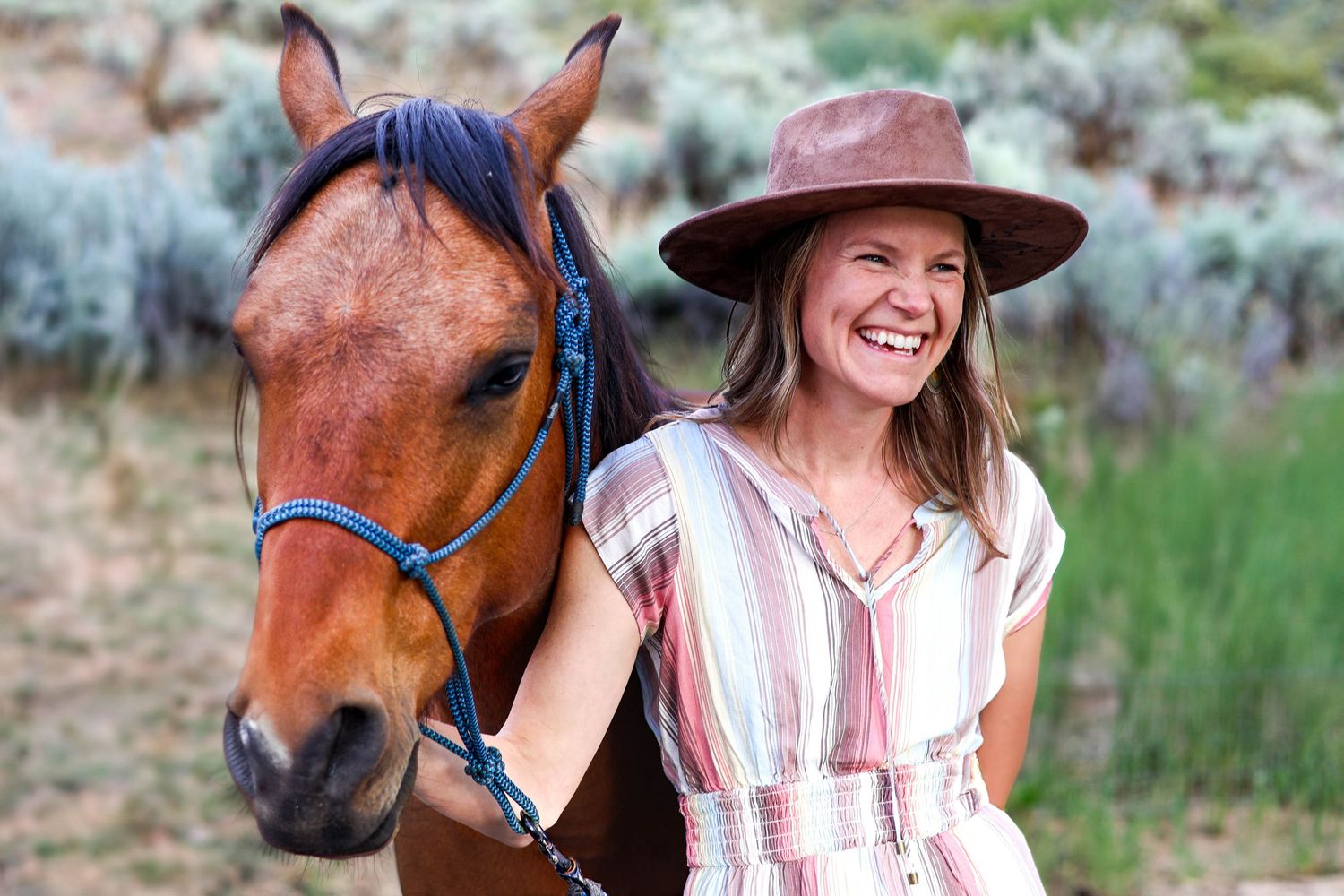 A person in a striped dress stands beside a brown horse wearing blue tack in a rural setting.