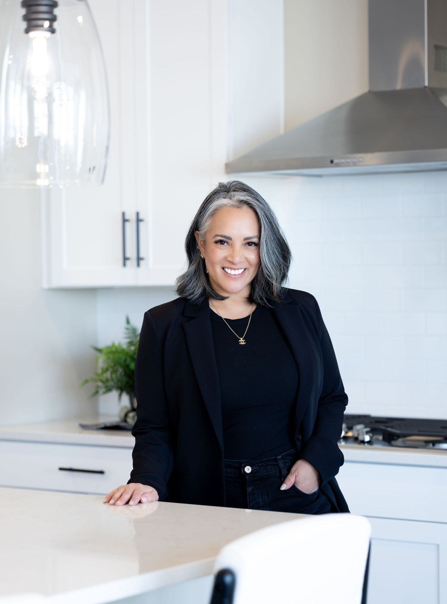 Professional woman in black blazer smiling while working at modern white kitchen counter with laptop.