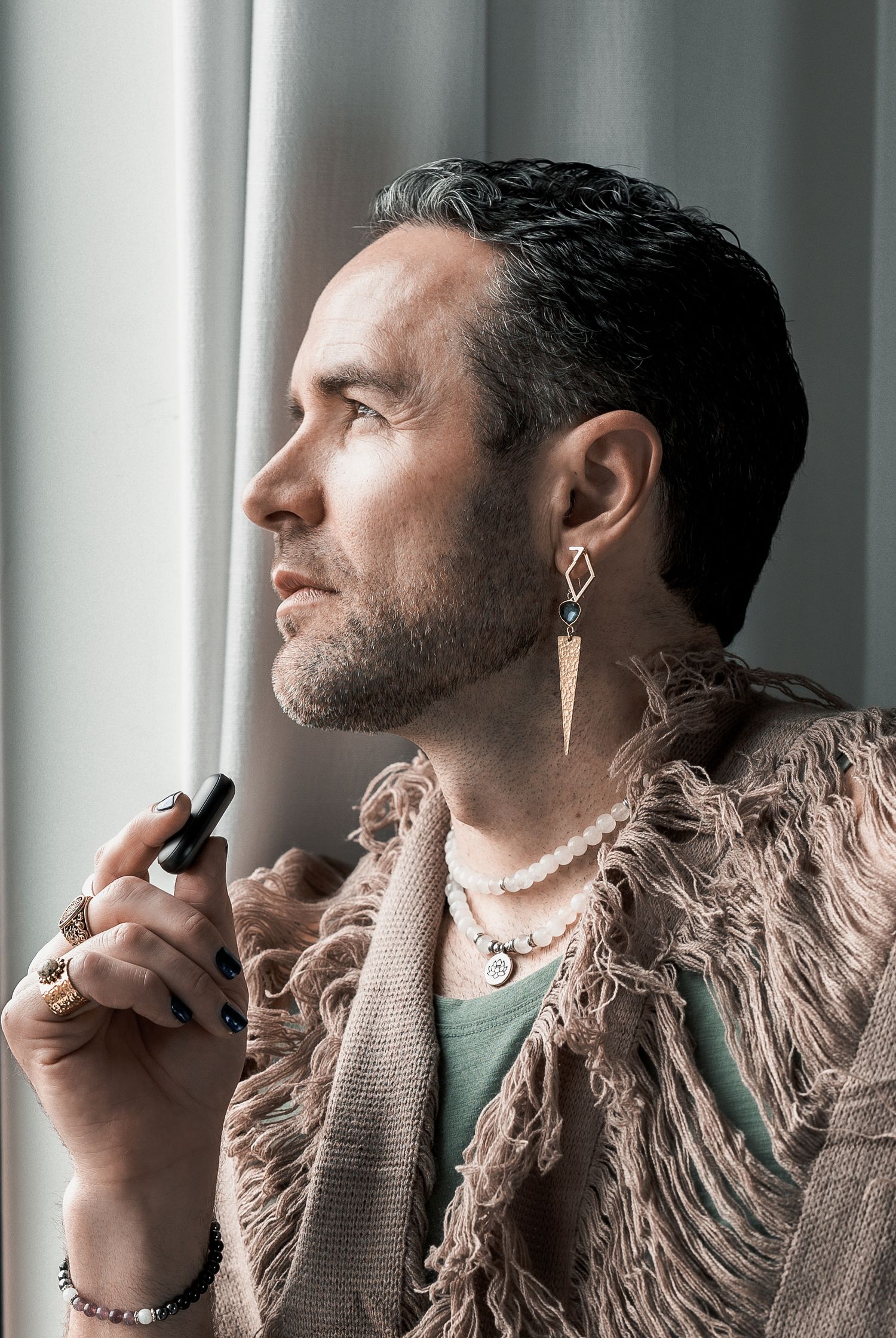 Profile view of person in beige fringe cardigan with earrings holding vape device near window in moody lighting.