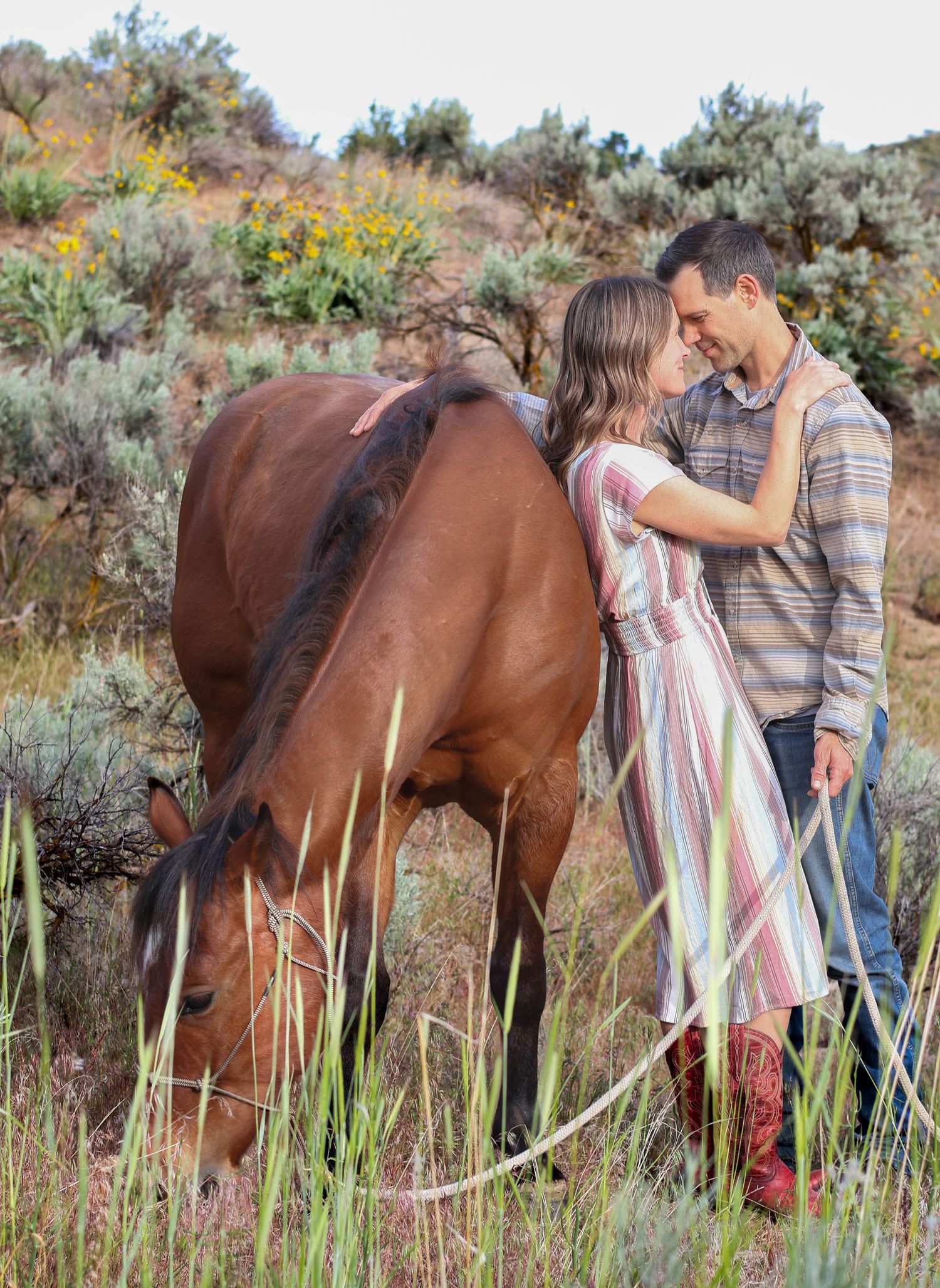 A couple embraces near their grazing brown horse among sagebrush and wildflowers.