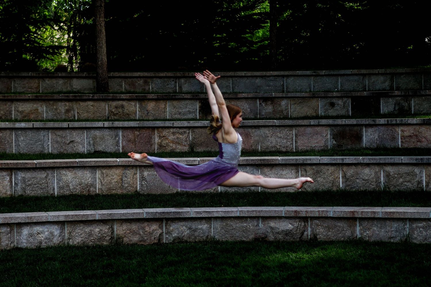A dancer in a purple leotard performing a graceful leap pose on stone steps outdoors.