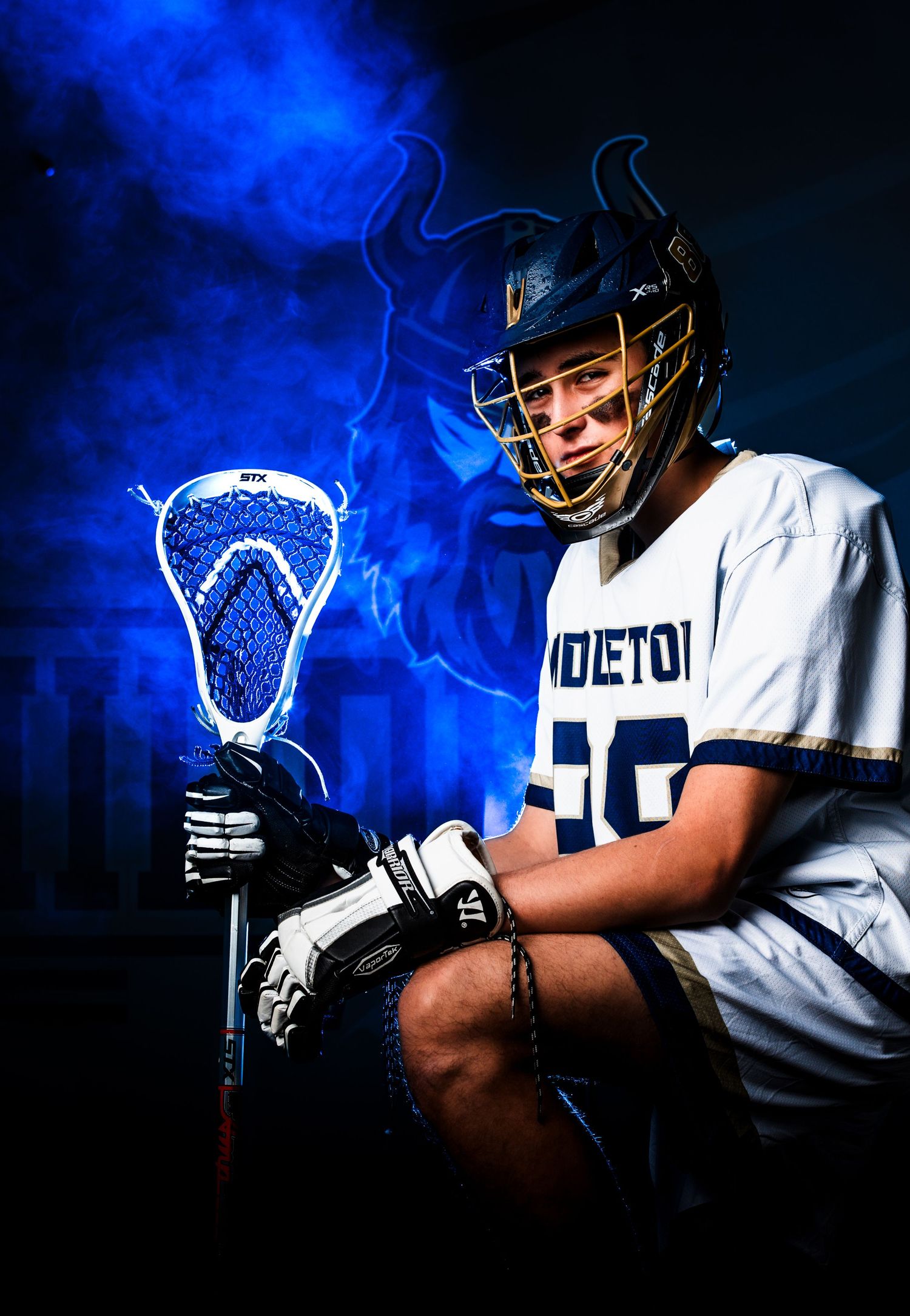 Lacrosse player poses with glowing blue stick against dramatic dark background with smoke effects.