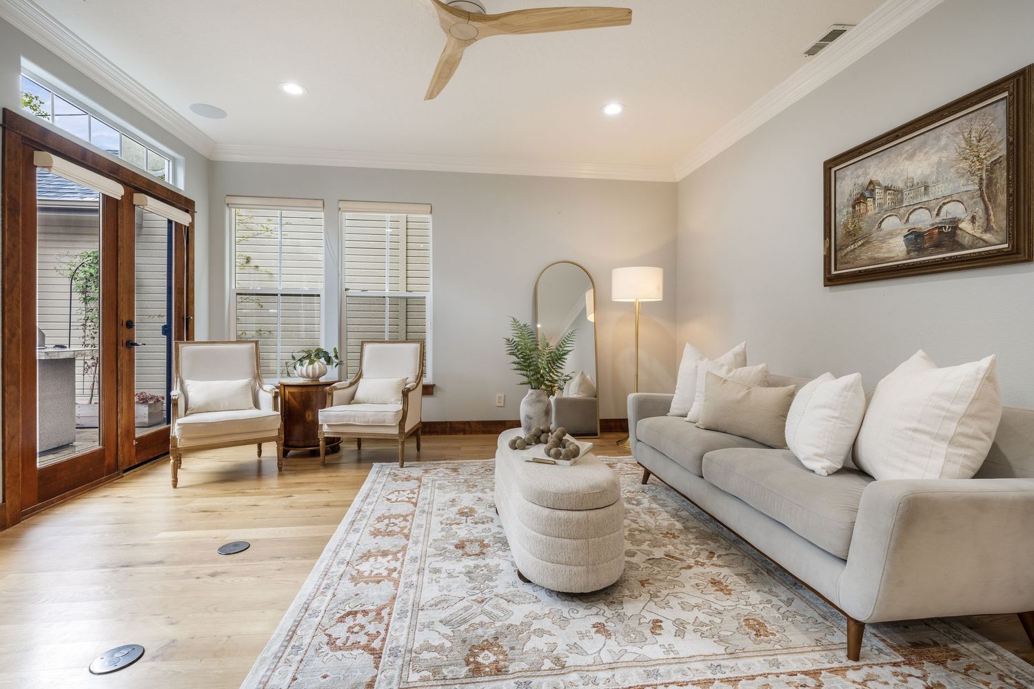 Bright living room with gray sofa, white armchairs, hardwood floors, ceiling fan, and French doors leading to outdoor space.