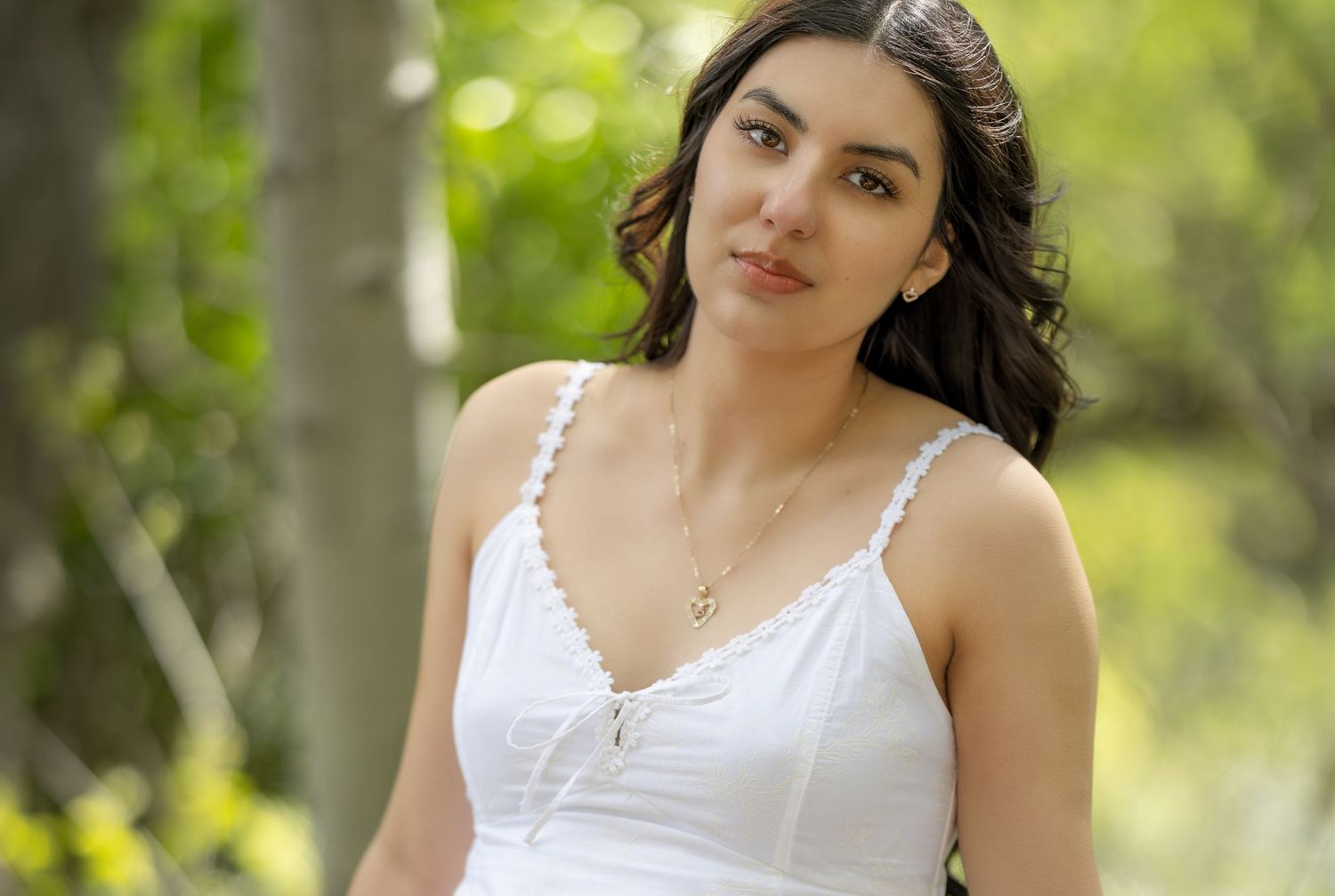 Young woman in white sundress with gold necklace posing outdoors among green trees in natural light.
