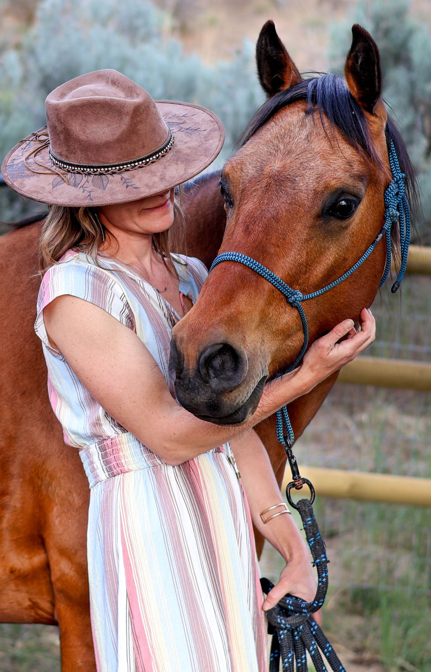 Person in brown hat and striped dress affectionately pets a brown horse wearing blue halter.