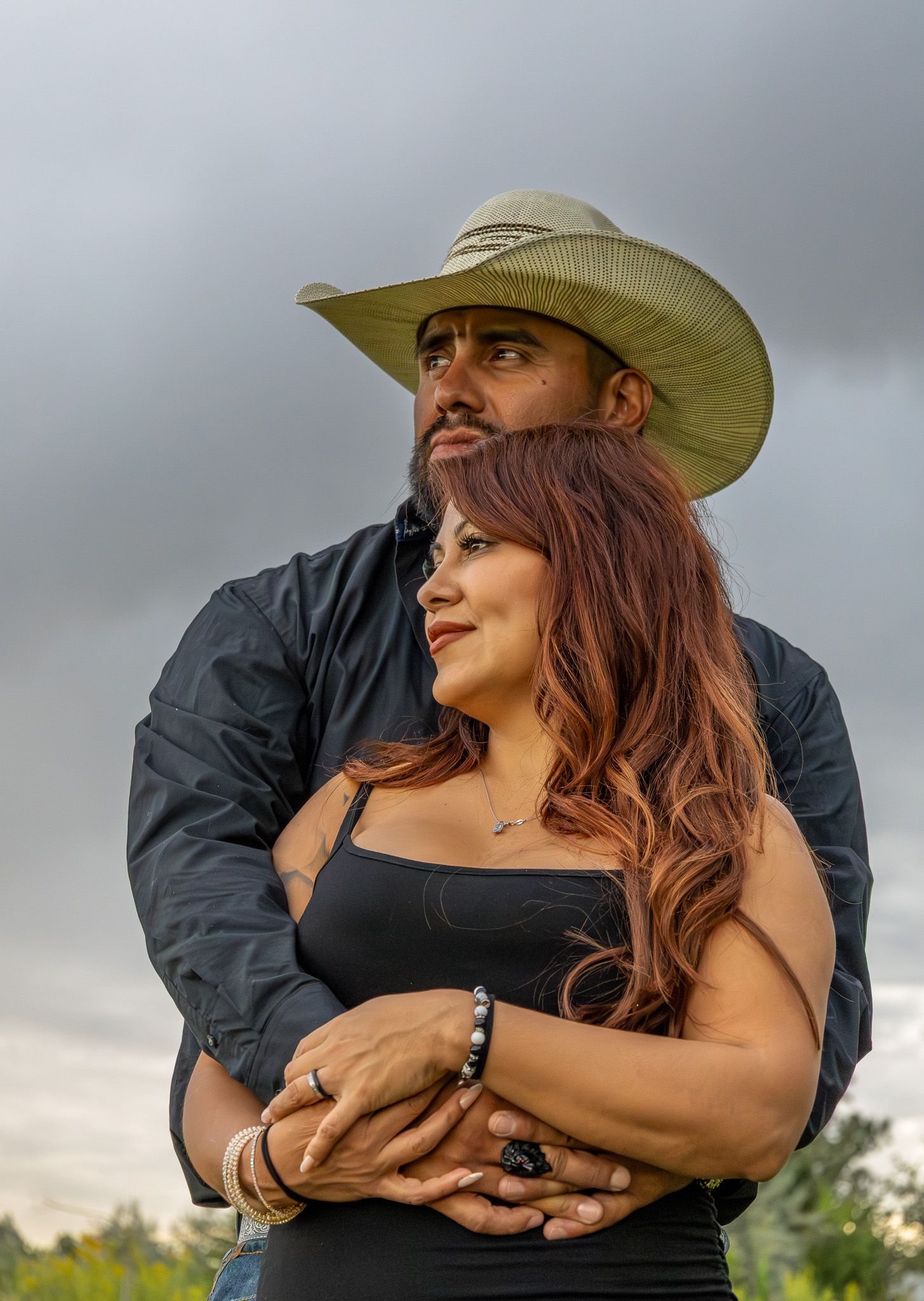 A romantic couple embraces outdoors wearing western attire against a cloudy sky.
