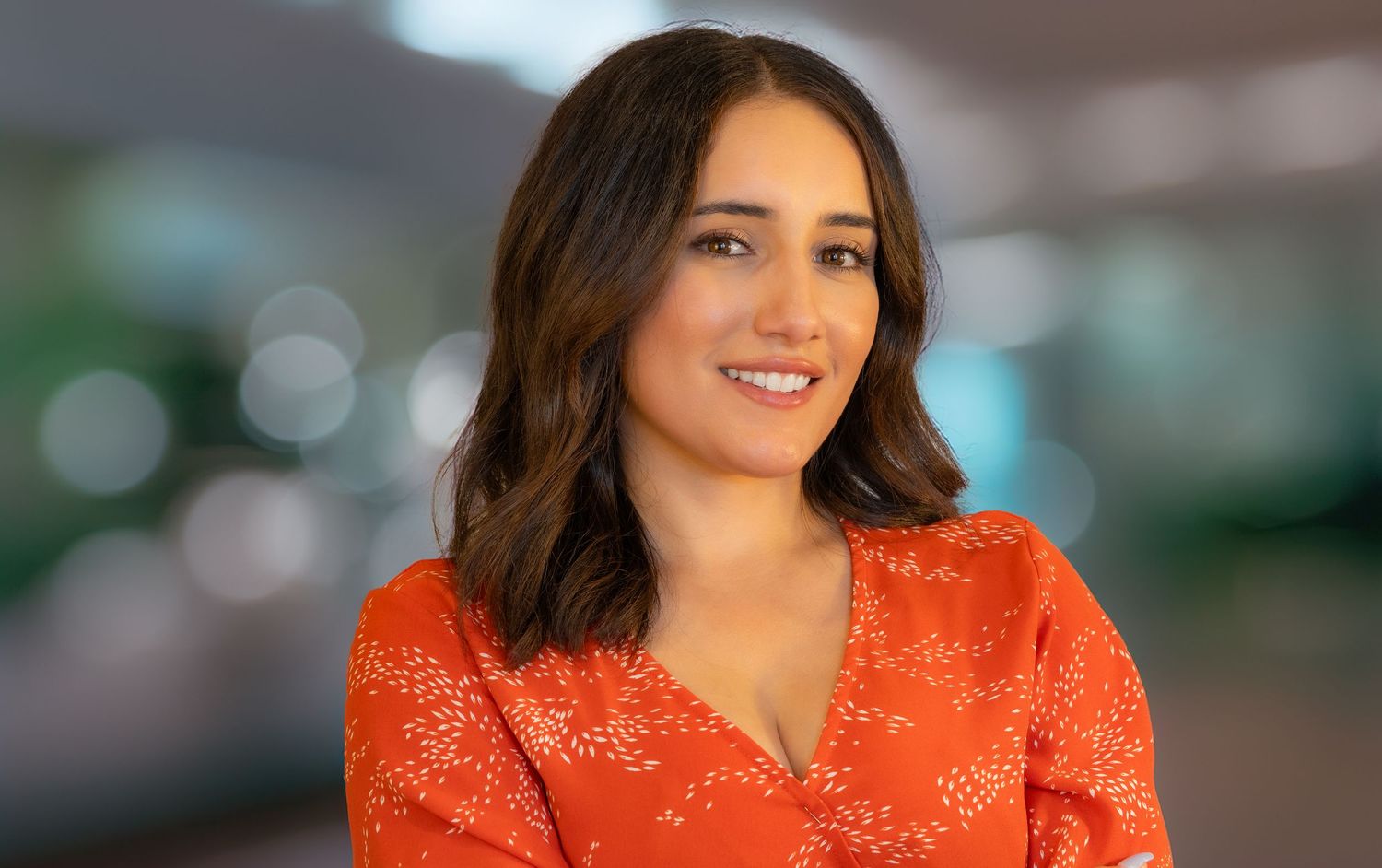 A person wearing a vibrant orange patterned blouse smiles warmly against a soft bokeh background.