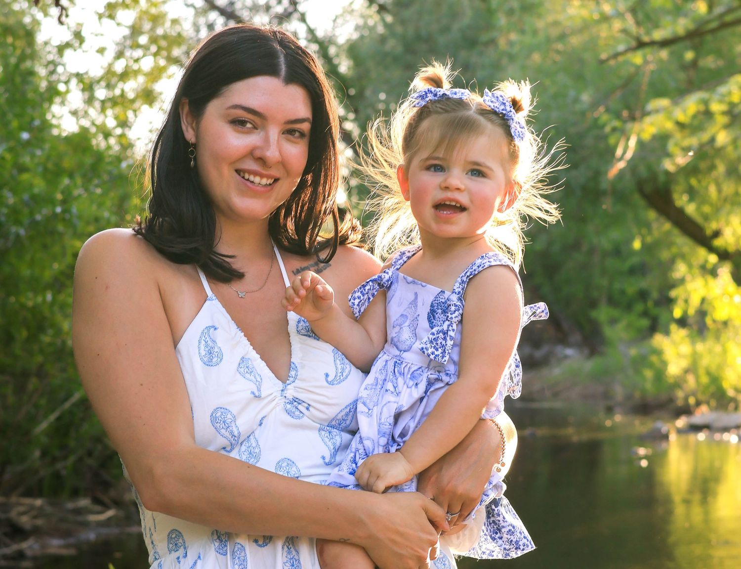 Two people wearing matching blue and white floral dresses pose together by a pond in a sunlit outdoor setting.