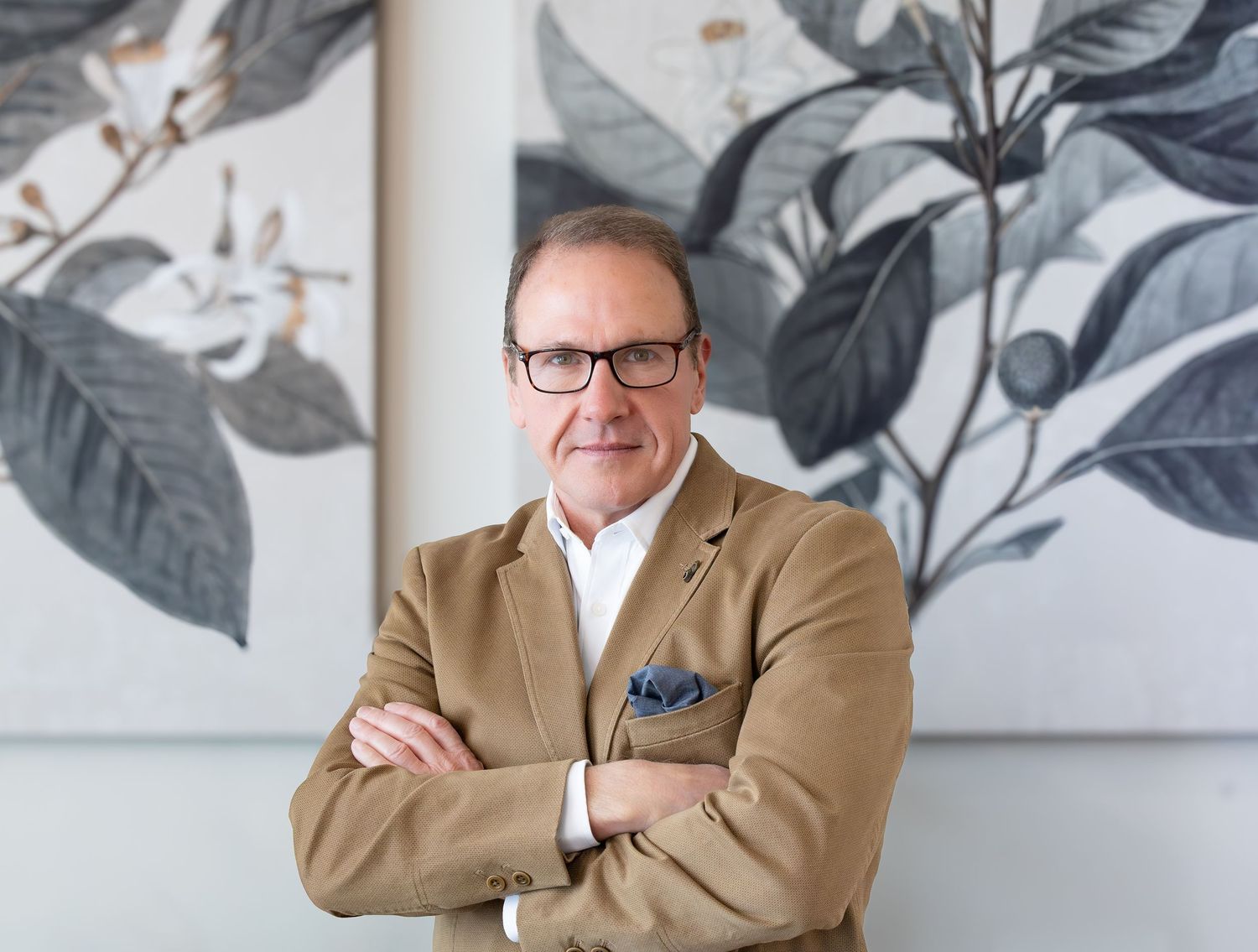 Professional wearing a tan blazer and glasses sits in front of botanical wall art in modern office setting.