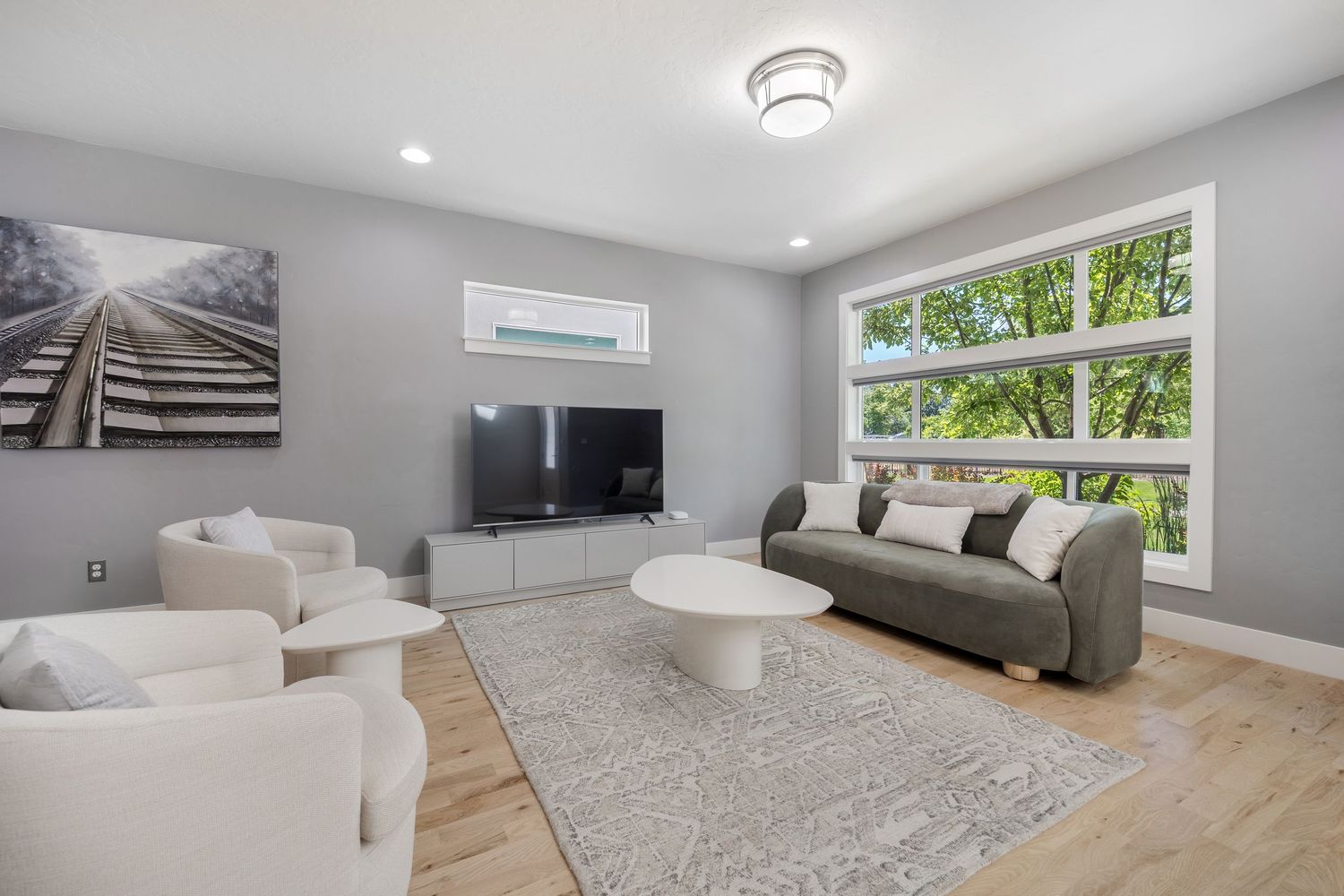 Modern living room with light wood floors, gray sofa, white chairs, round coffee table, large window, and wall-mounted TV.